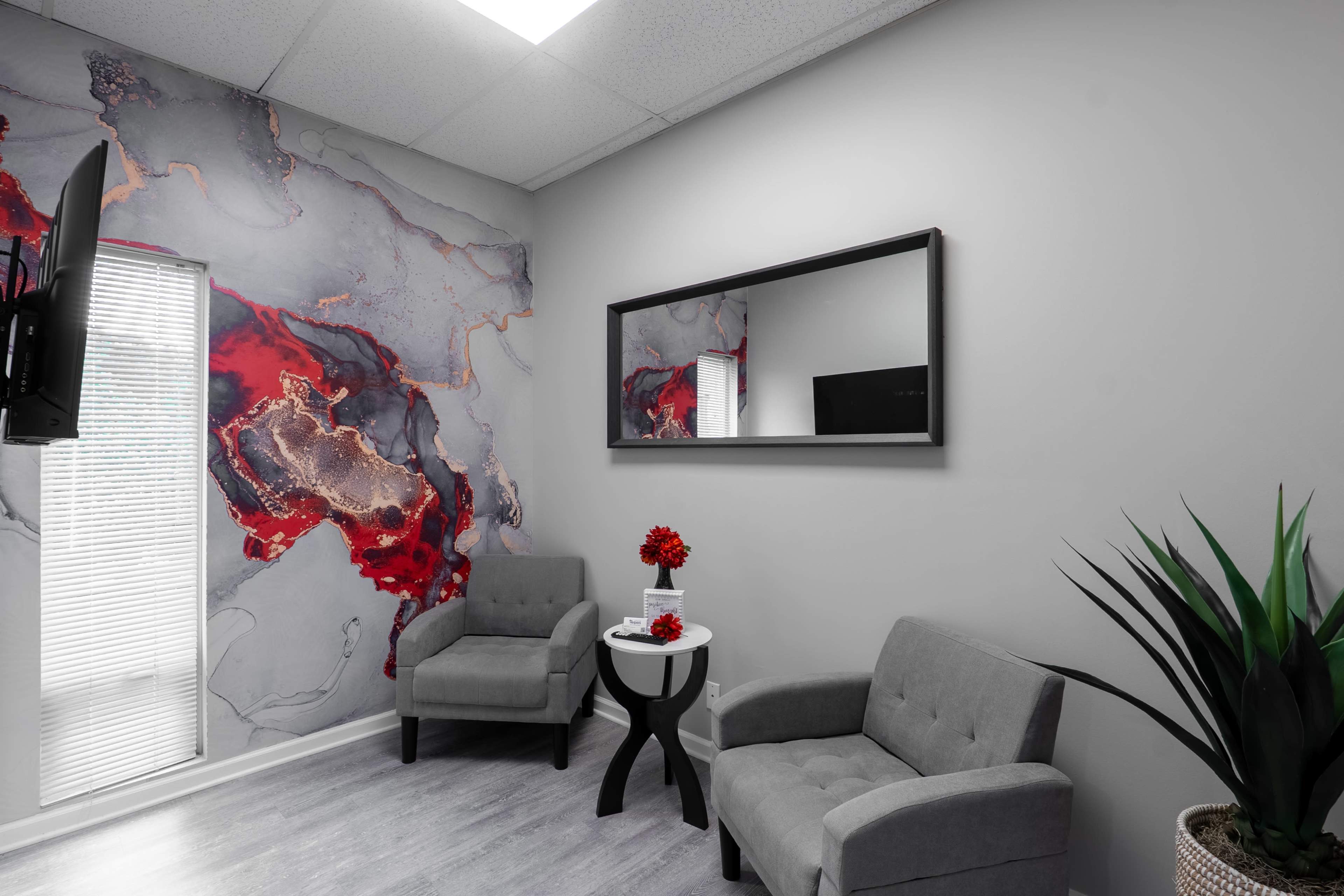 The image shows a modern waiting area with two gray chairs, a small round table with a flower arrangement, and a wall featuring a colorful abstract mural.