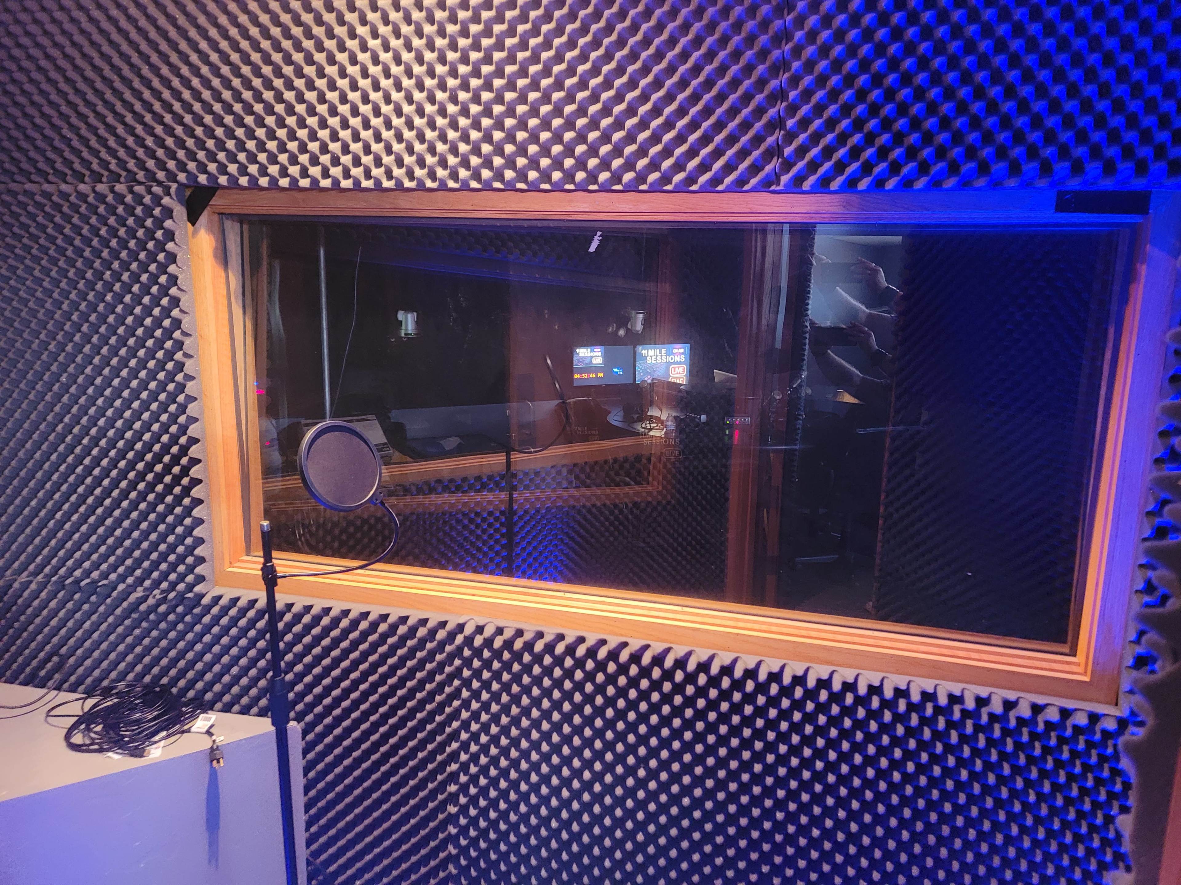 A recording studio booth with soundproof walls and a microphone in front of a glass window looking into a control room.