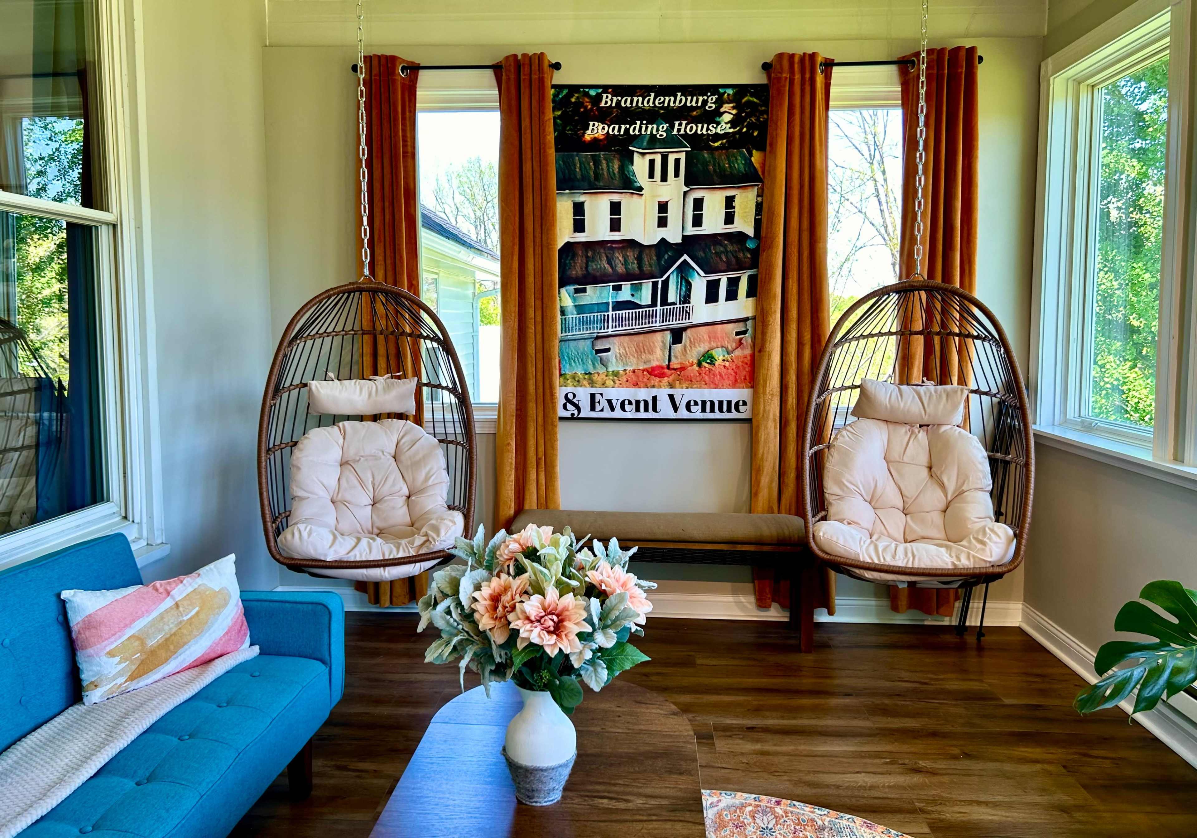 The image shows a brightly lit room featuring two hanging chairs, a blue couch, a decorative table, and a large banner for Brandenburg Boarding House & Event Venue on the wall.