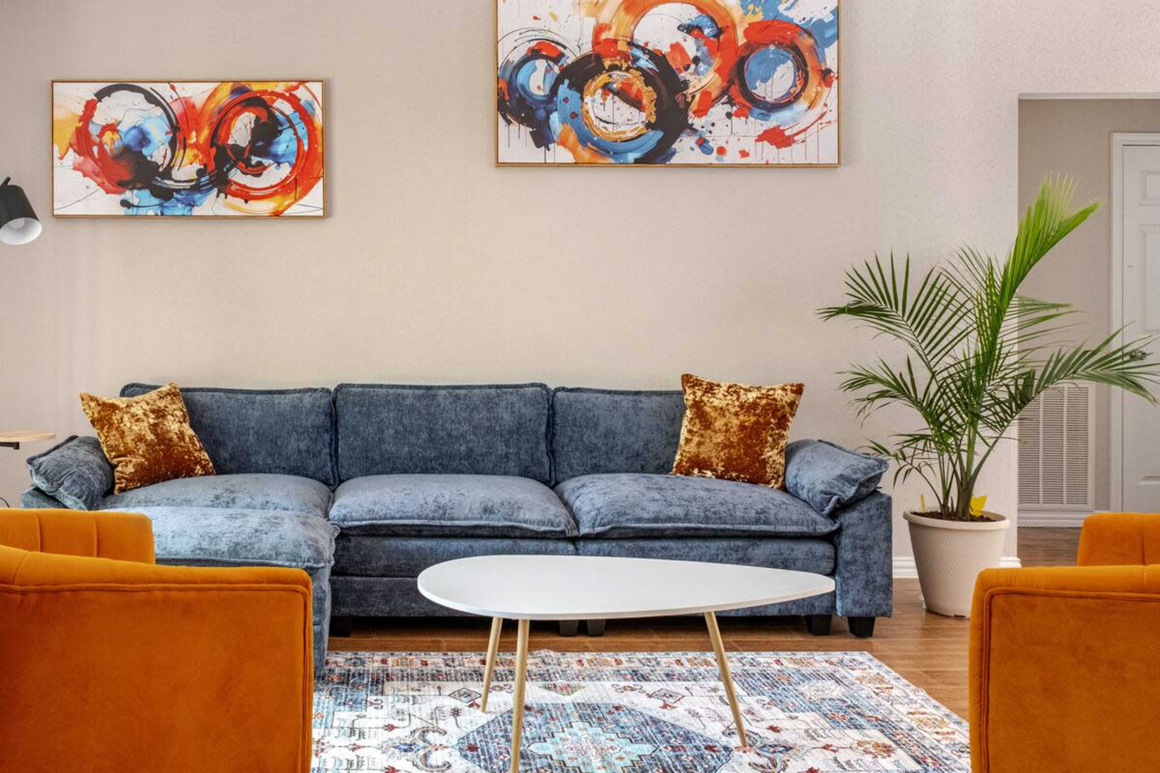 The image shows a living room with a blue couch, orange accent chairs, a white coffee table, and colorful abstract paintings on the walls.