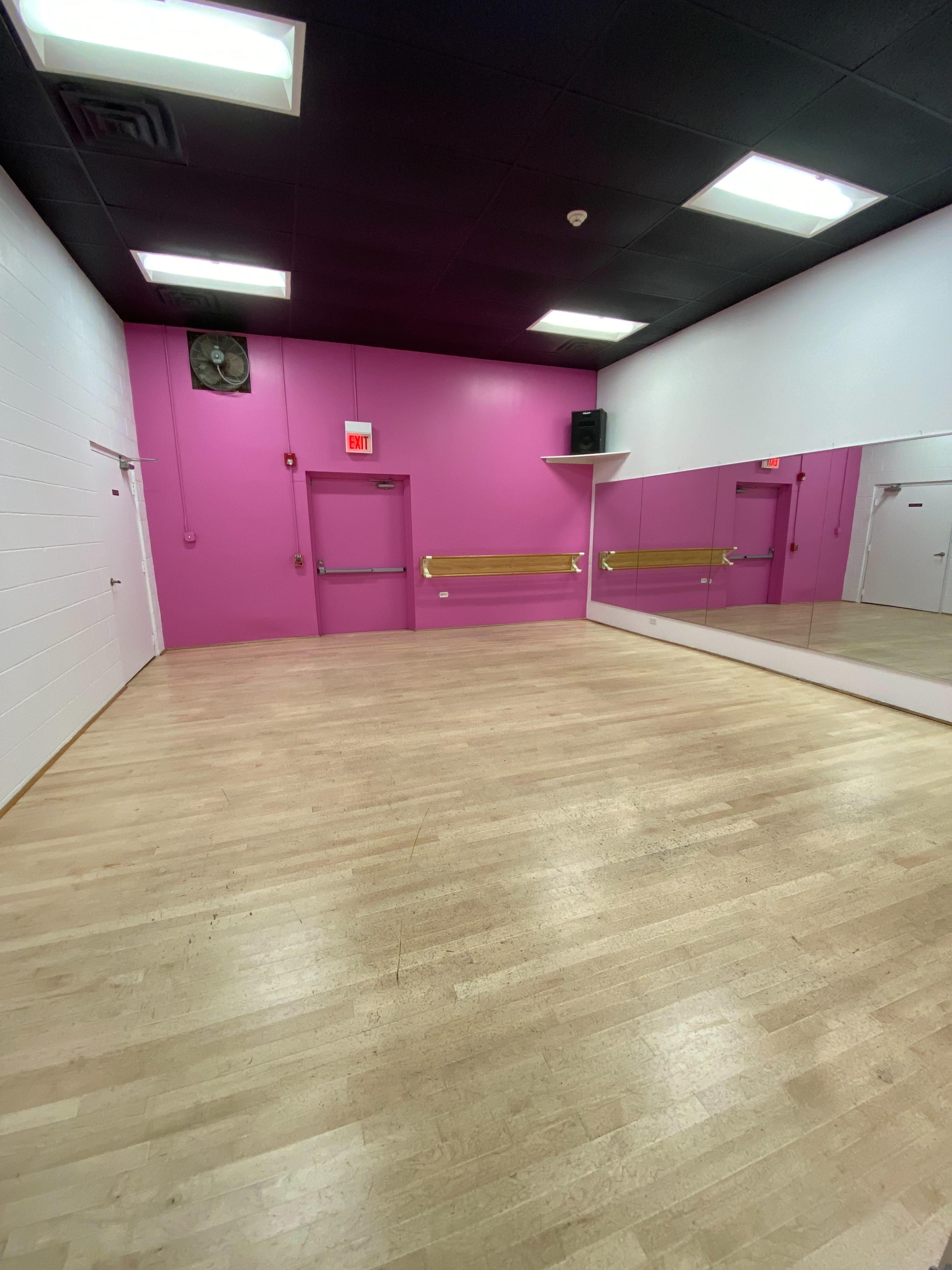 A dance studio with light wood flooring, pink walls, and a large mirror on one side.