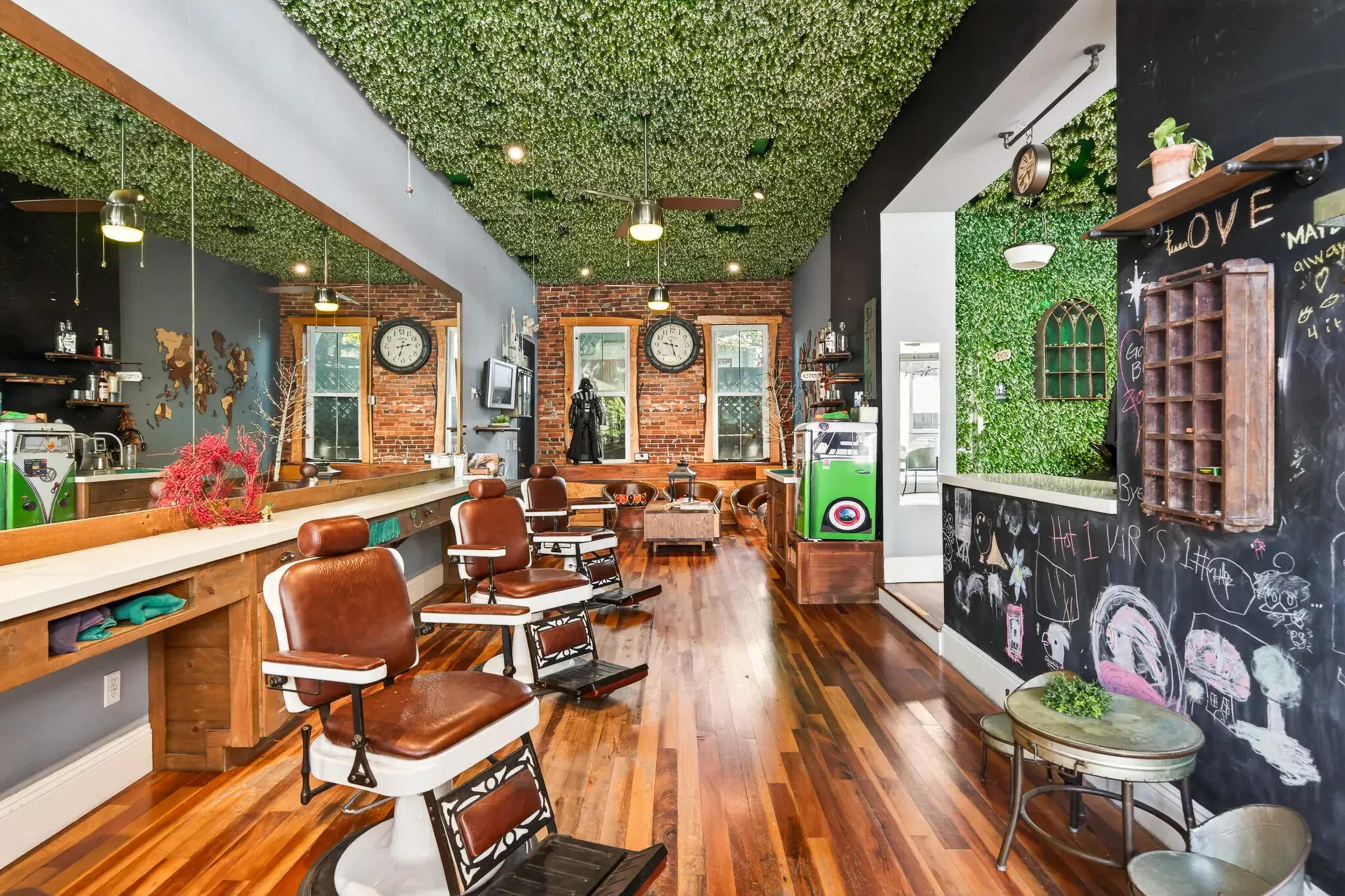 A hair salon features brown chairs, wooden flooring, and a green wall with plants, complemented by large windows.