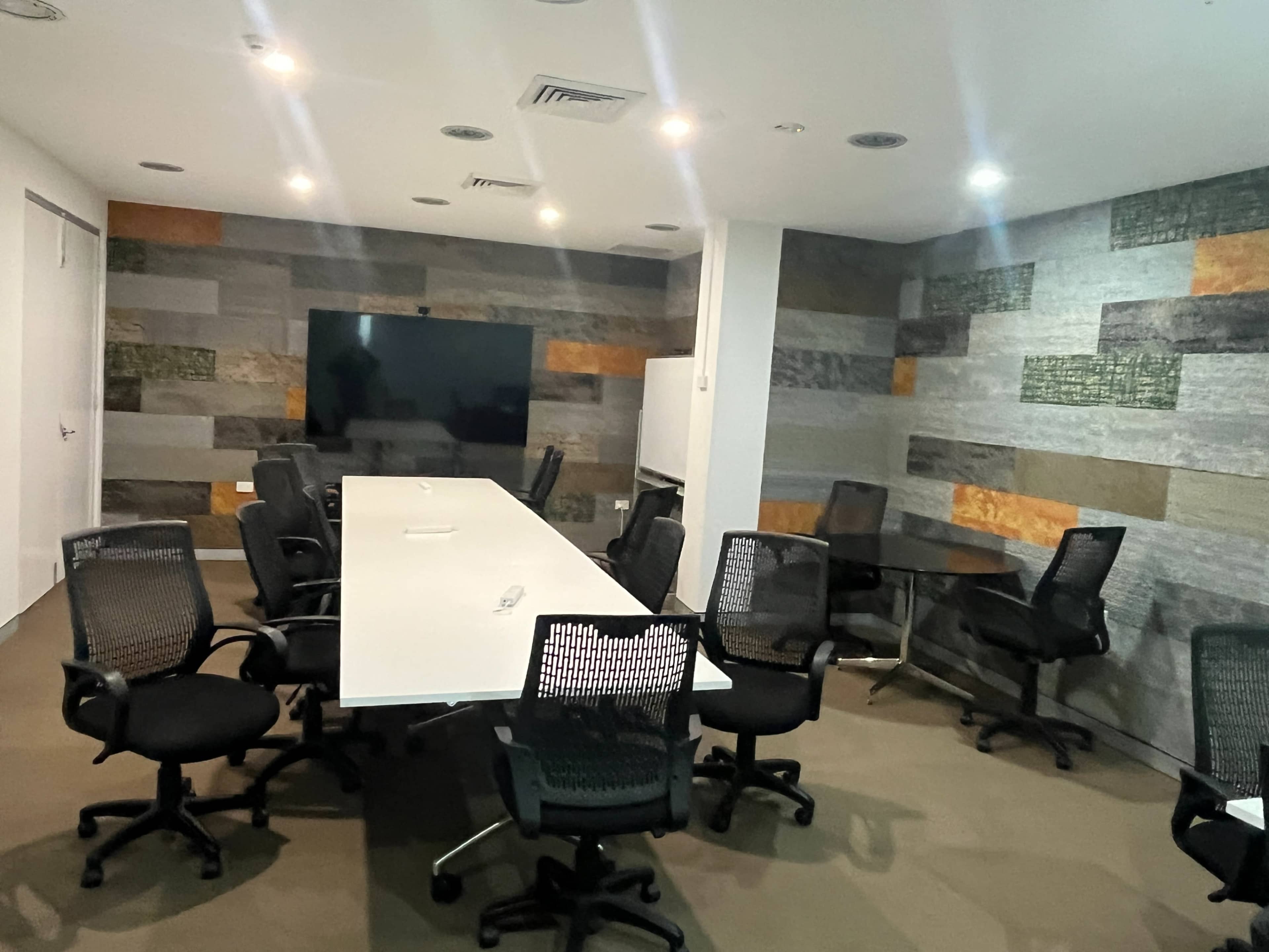 A modern conference room features a long white table surrounded by black mesh chairs, with a large screen on one wall and textured panels on the others.