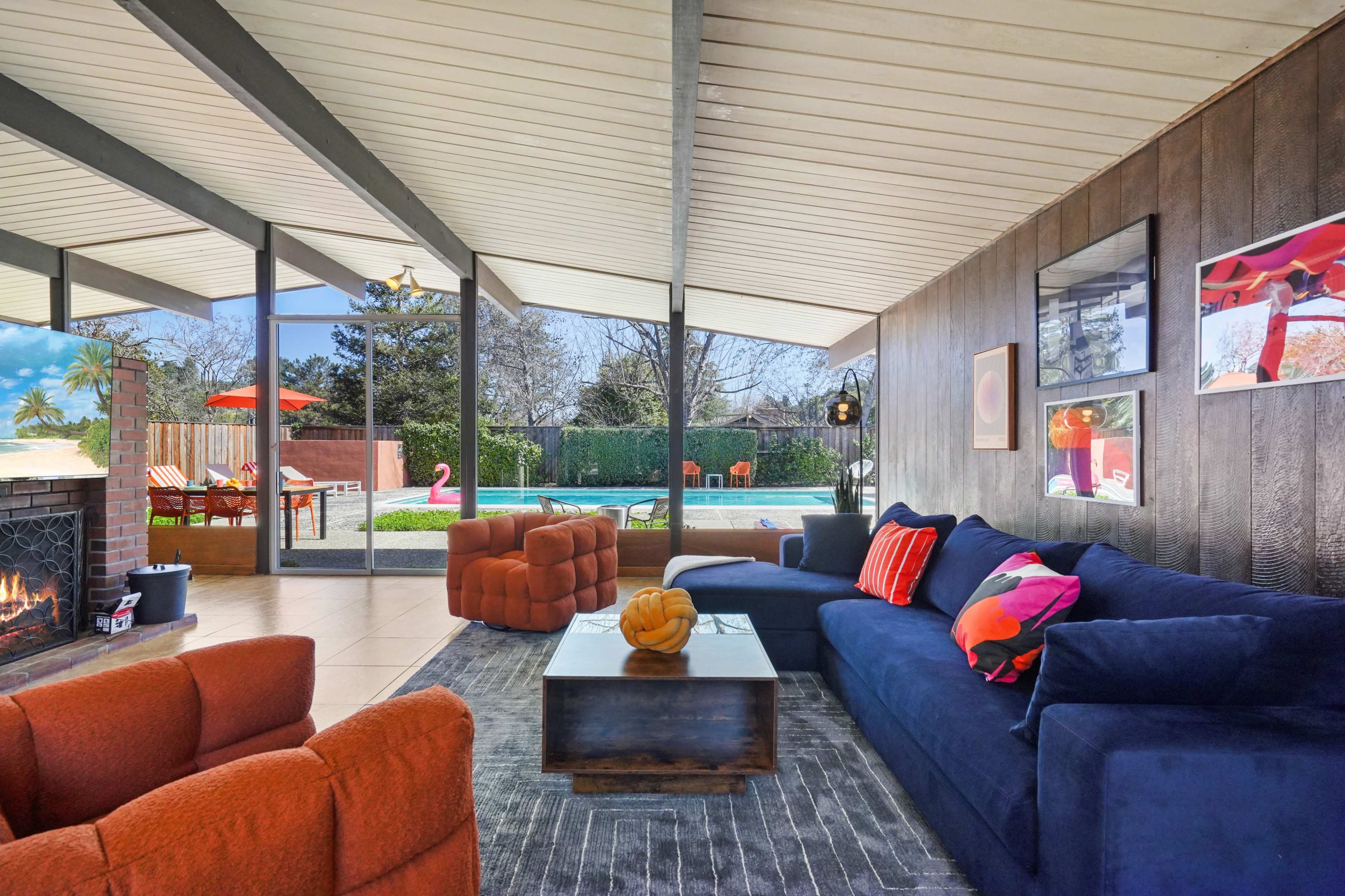 The image shows a modern living room with large glass windows overlooking a backyard pool area, featuring colorful furniture and a fireplace.