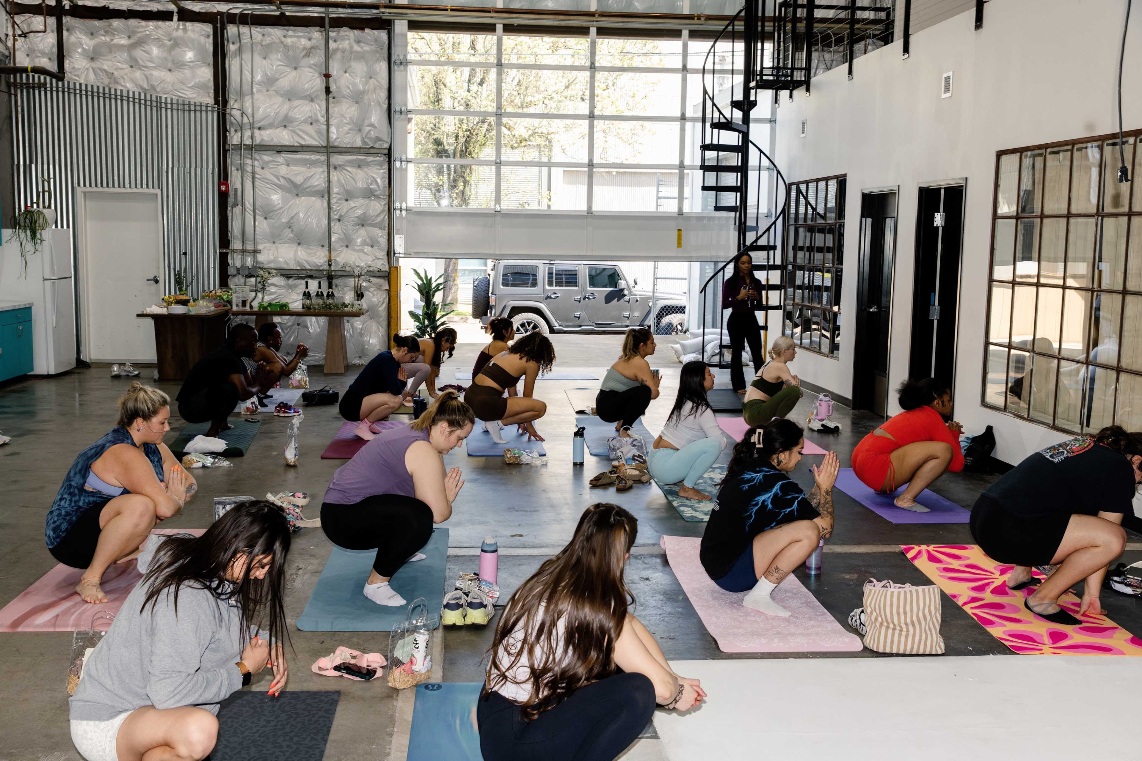 A group of people is practicing yoga in a spacious studio with large windows and natural light.
