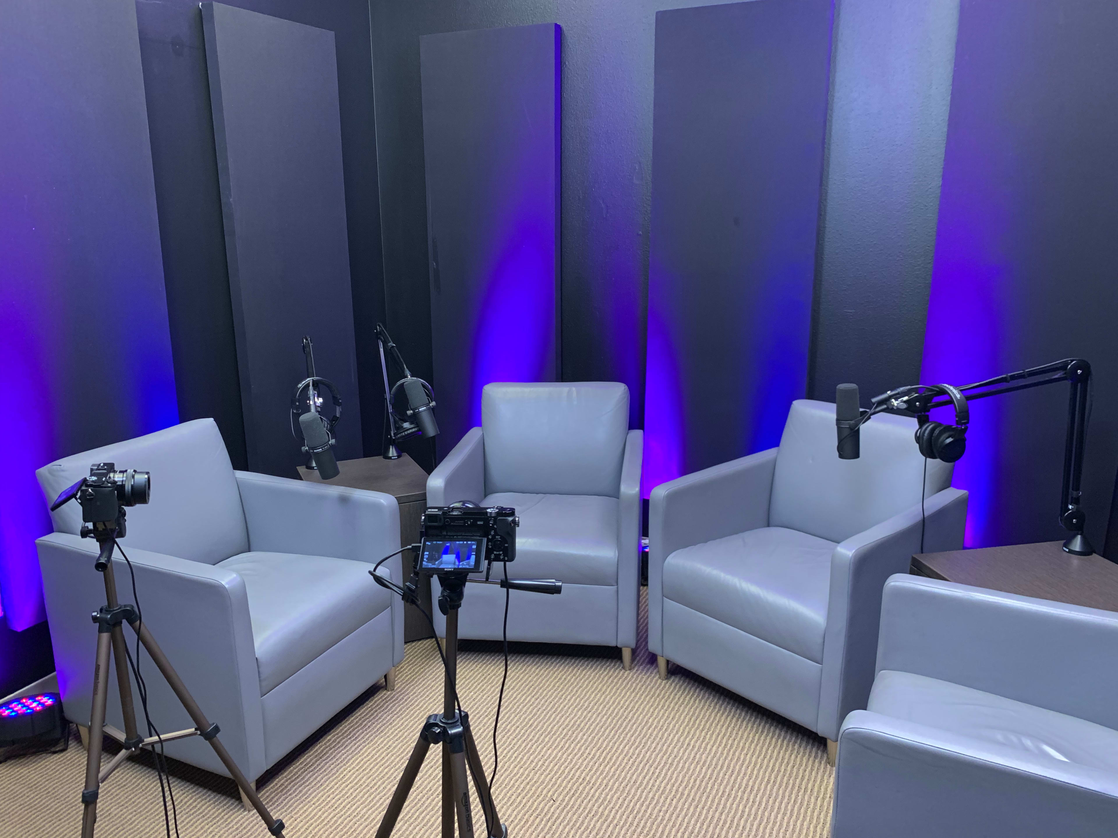 A small studio setup featuring three gray chairs arranged around a table, with cameras on tripods and microphones positioned for recording.