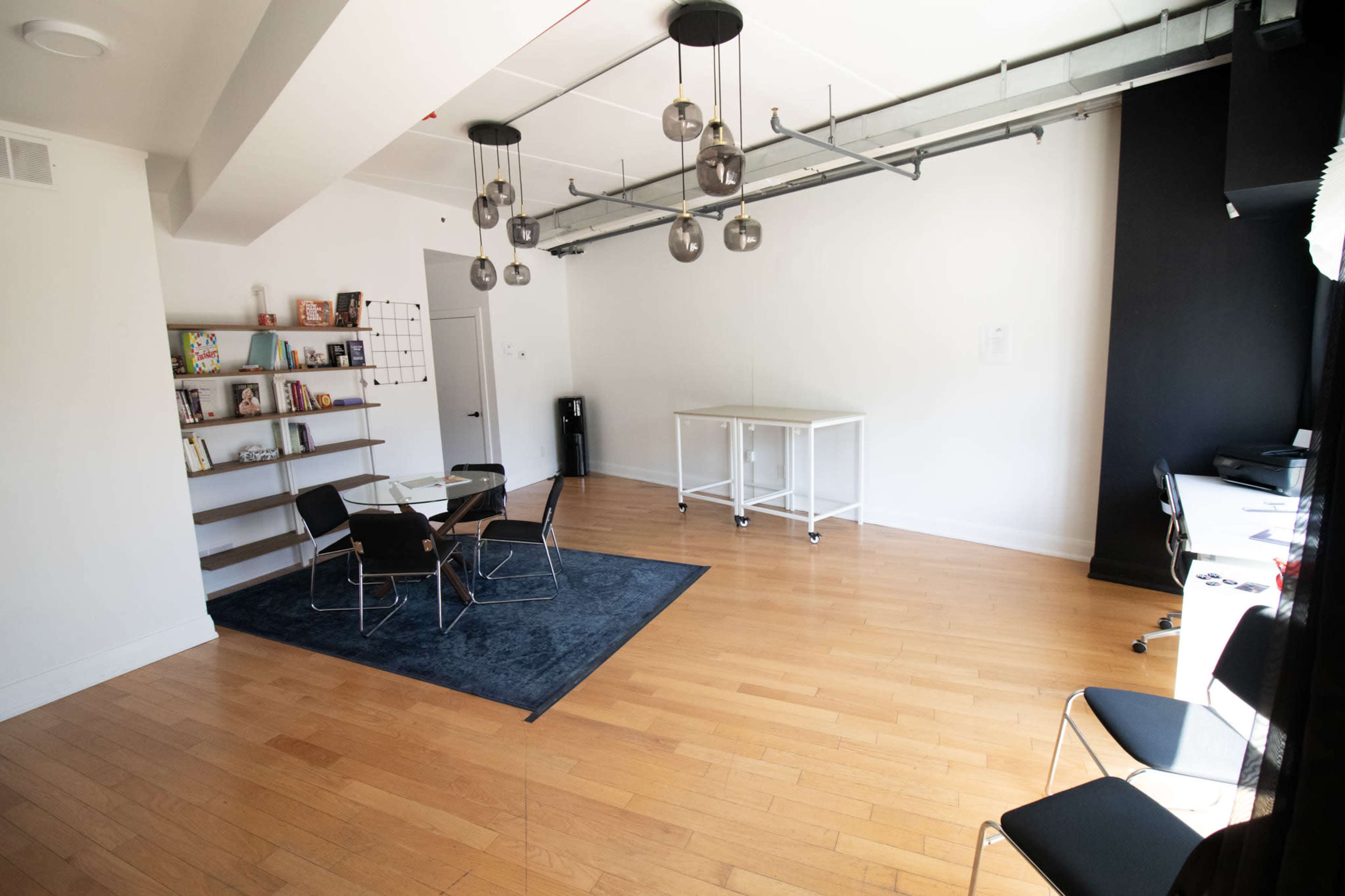 The image shows a spacious, minimally furnished room with hardwood floors, a black accent wall, a glass meeting table surrounded by chairs, and shelving displaying books.