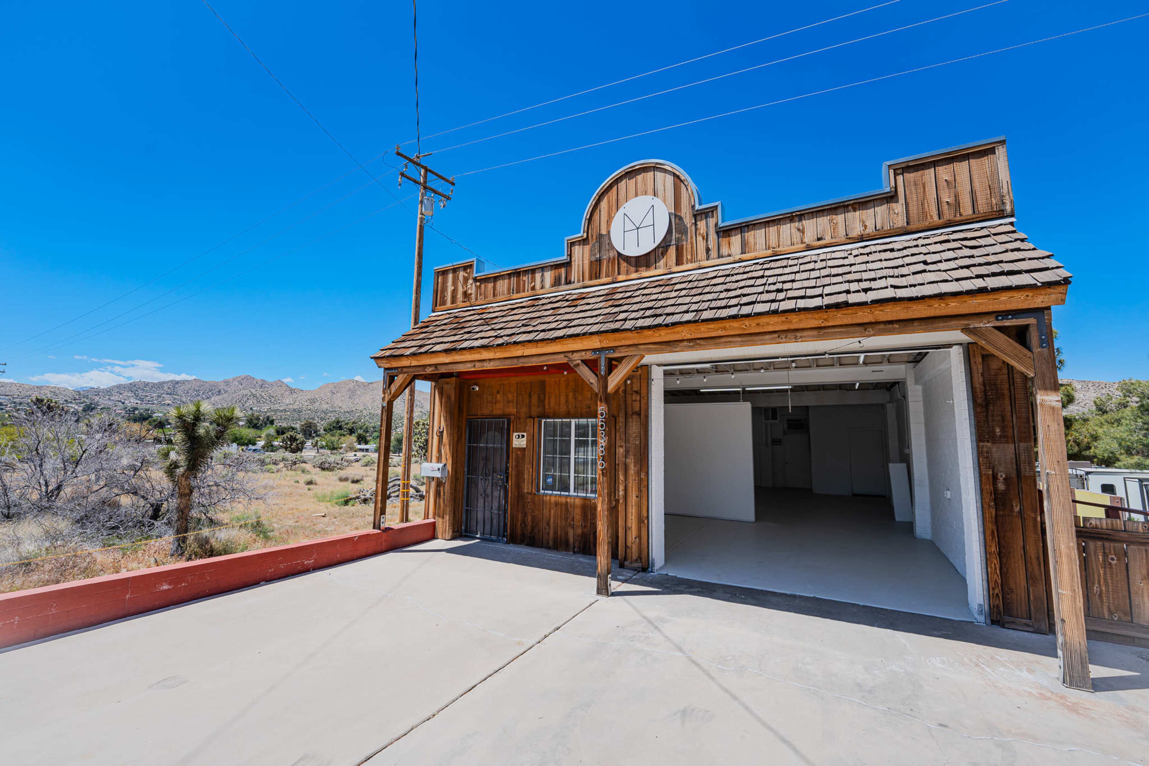 A wooden structure with a gabled roof and circular emblem stands in a desert landscape, revealing an interior space with an open layout.