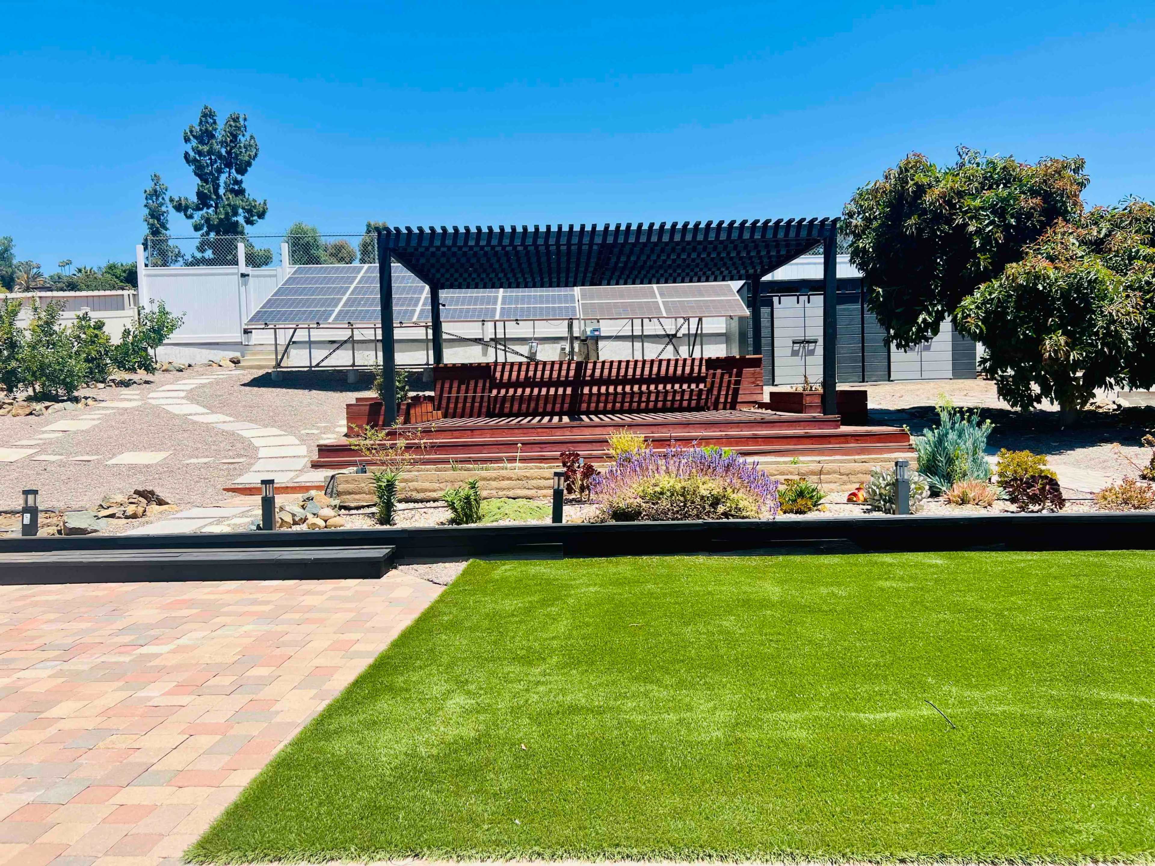 The image shows a landscaped backyard with a pergola, flower beds, and solar panels in the background.