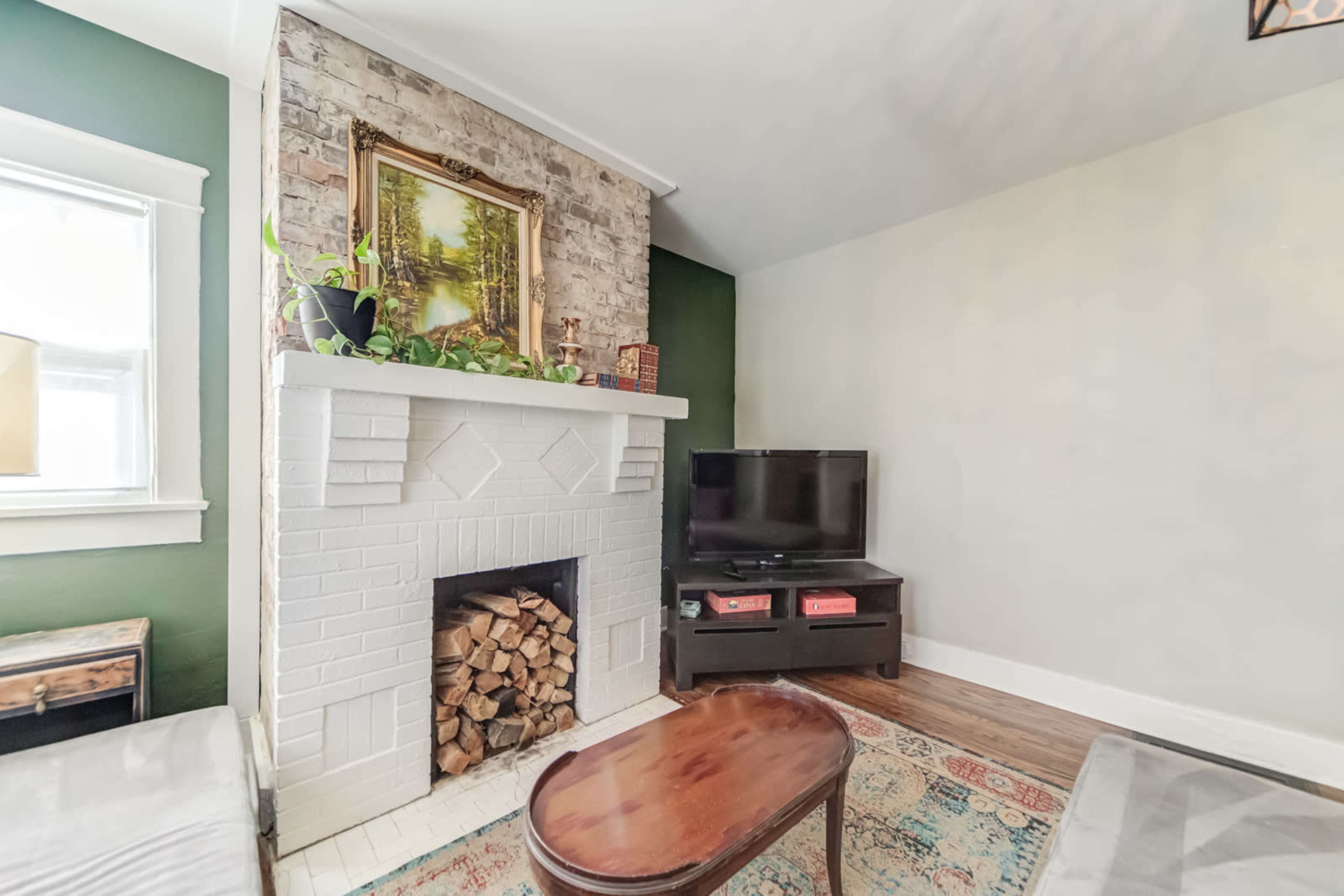 A living room with a white fireplace stacked with firewood, a wooden coffee table, and a TV on a black stand, against a backdrop of a green wall and exposed brick.