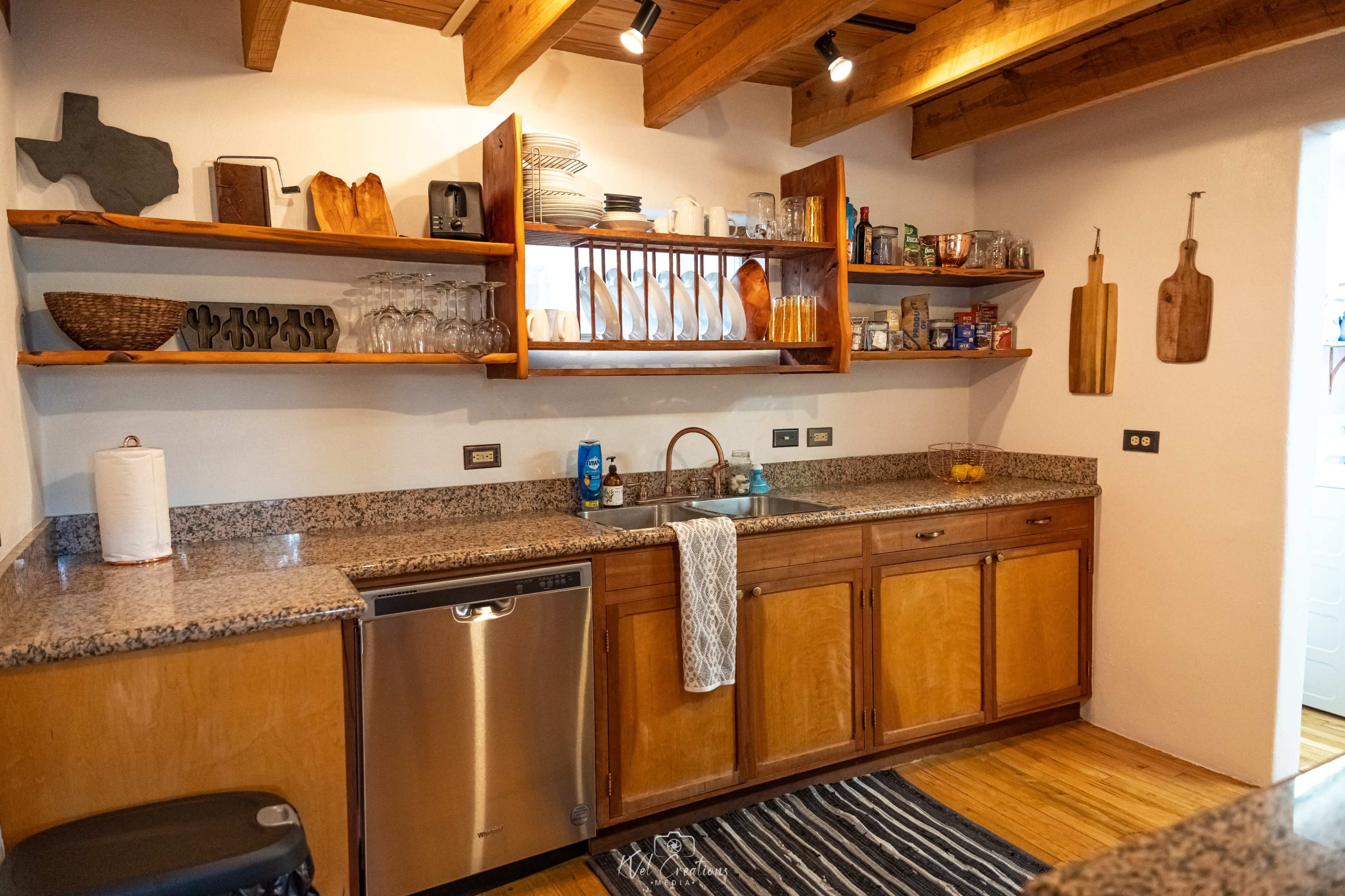 The image shows a kitchen with wooden shelves displaying dishes and glasses, a granite countertop, and appliances including a dishwasher.