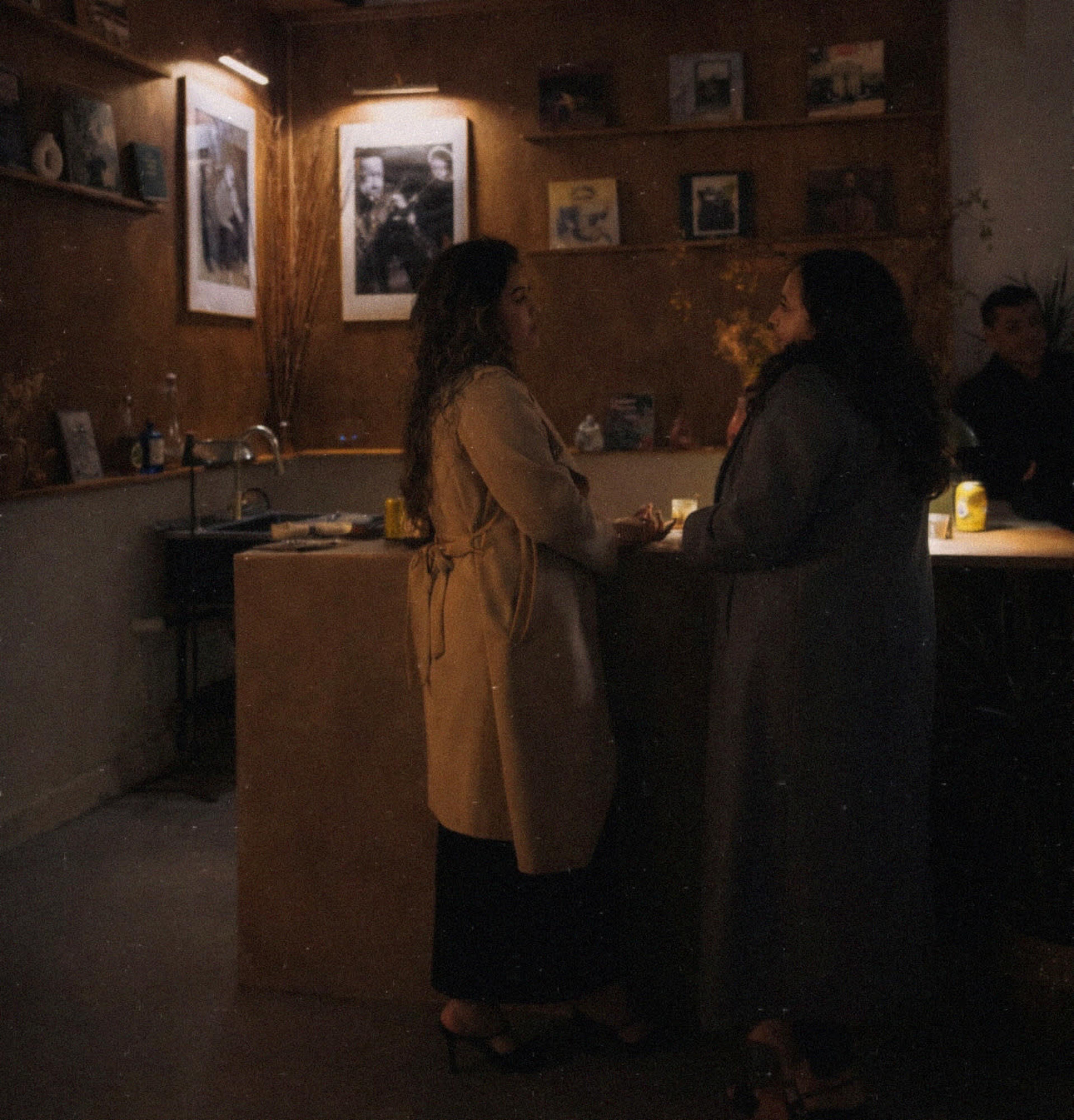 Two women in long coats are engaged in conversation at a bar in a dimly lit space adorned with framed photographs.