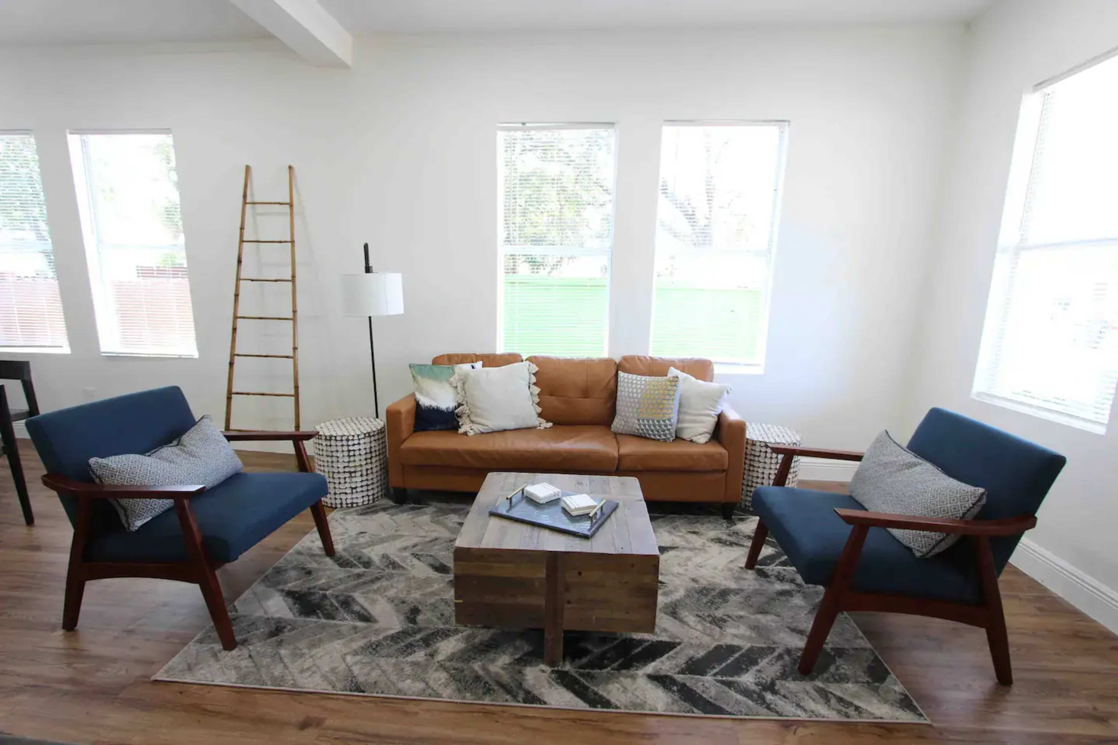 The scene features a modern living room with a brown leather sofa, two blue armchairs, a wooden coffee table, and large windows allowing natural light to enter.