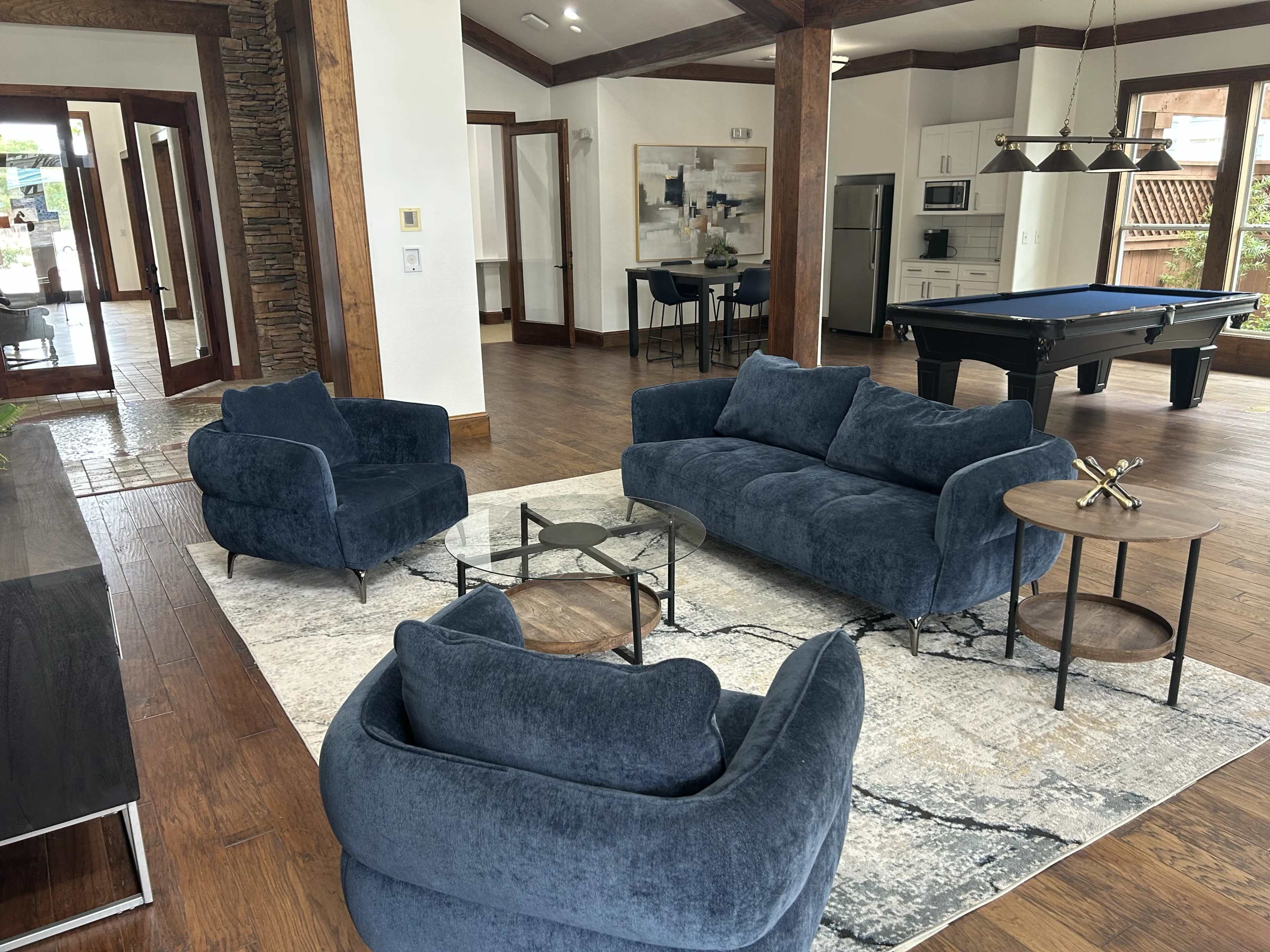 A modern living room features a blue sofa and armchairs arranged around a glass coffee table on a large area rug, with a kitchen and pool table visible in the background.