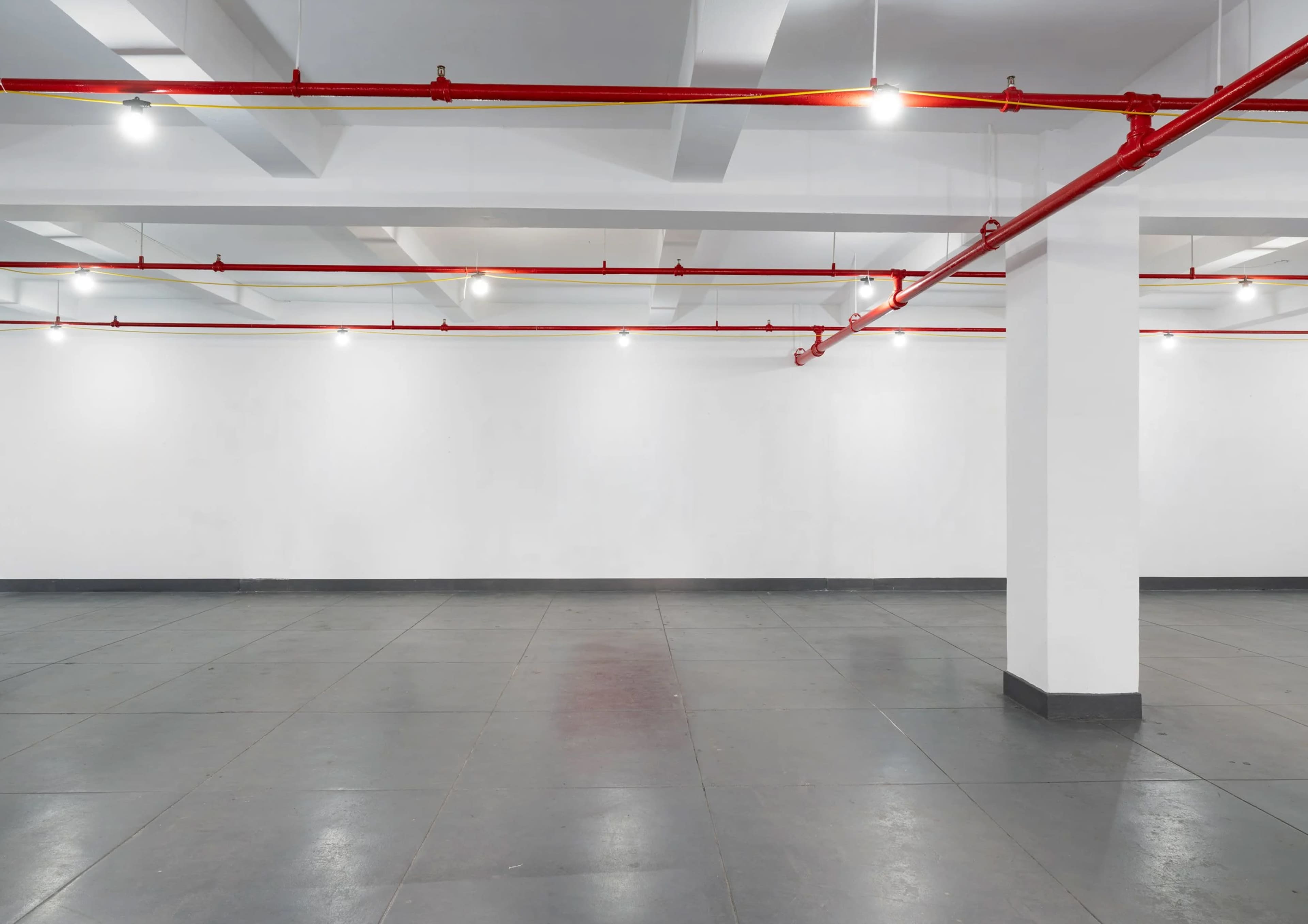 The image shows a spacious, empty room with a gray floor, white walls, and red pipes running along the ceiling.