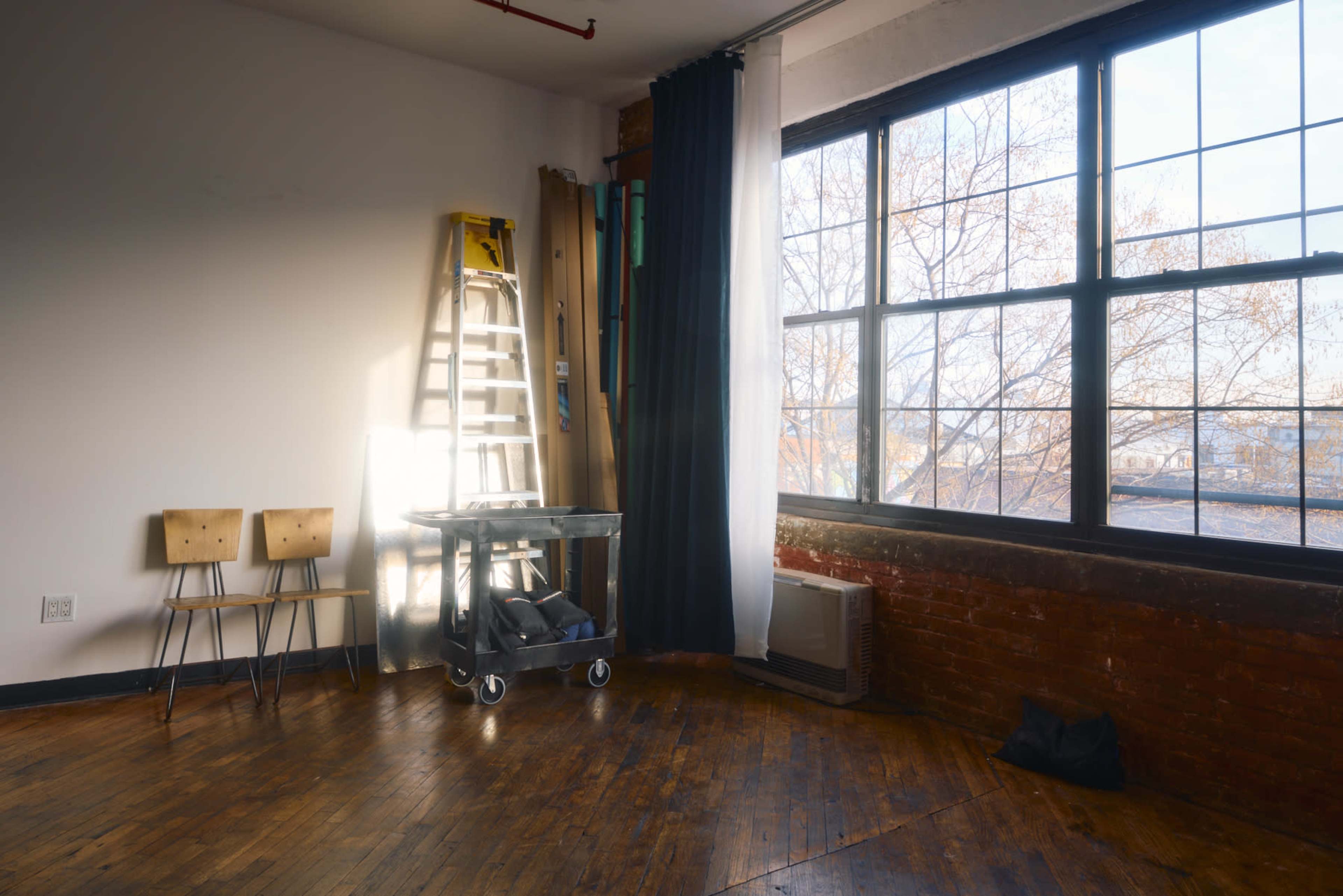 The image shows a well-lit room featuring two wooden chairs, a black cart, a ladder, and large windows with visible tree branches outside.