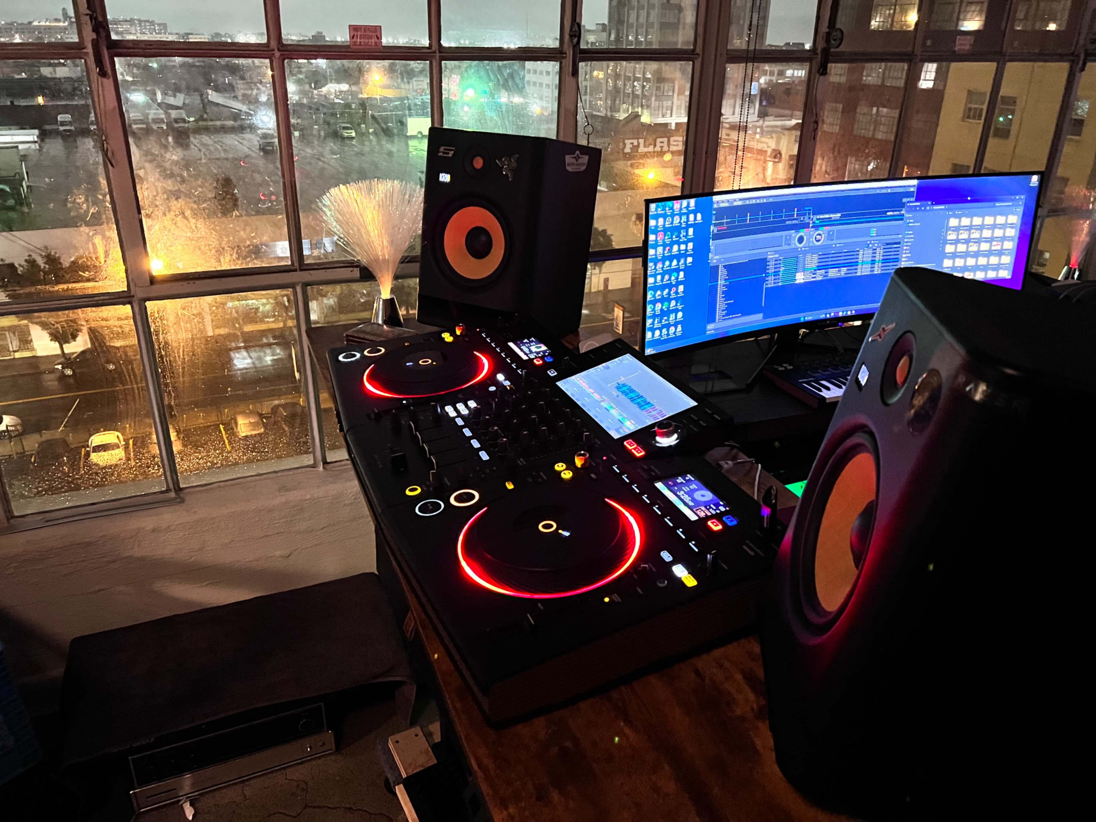 The image shows a DJ setup featuring a mixer and turntables with glowing lights, alongside two speakers and a computer displaying music software, all viewed through a large window with rain visible outside.