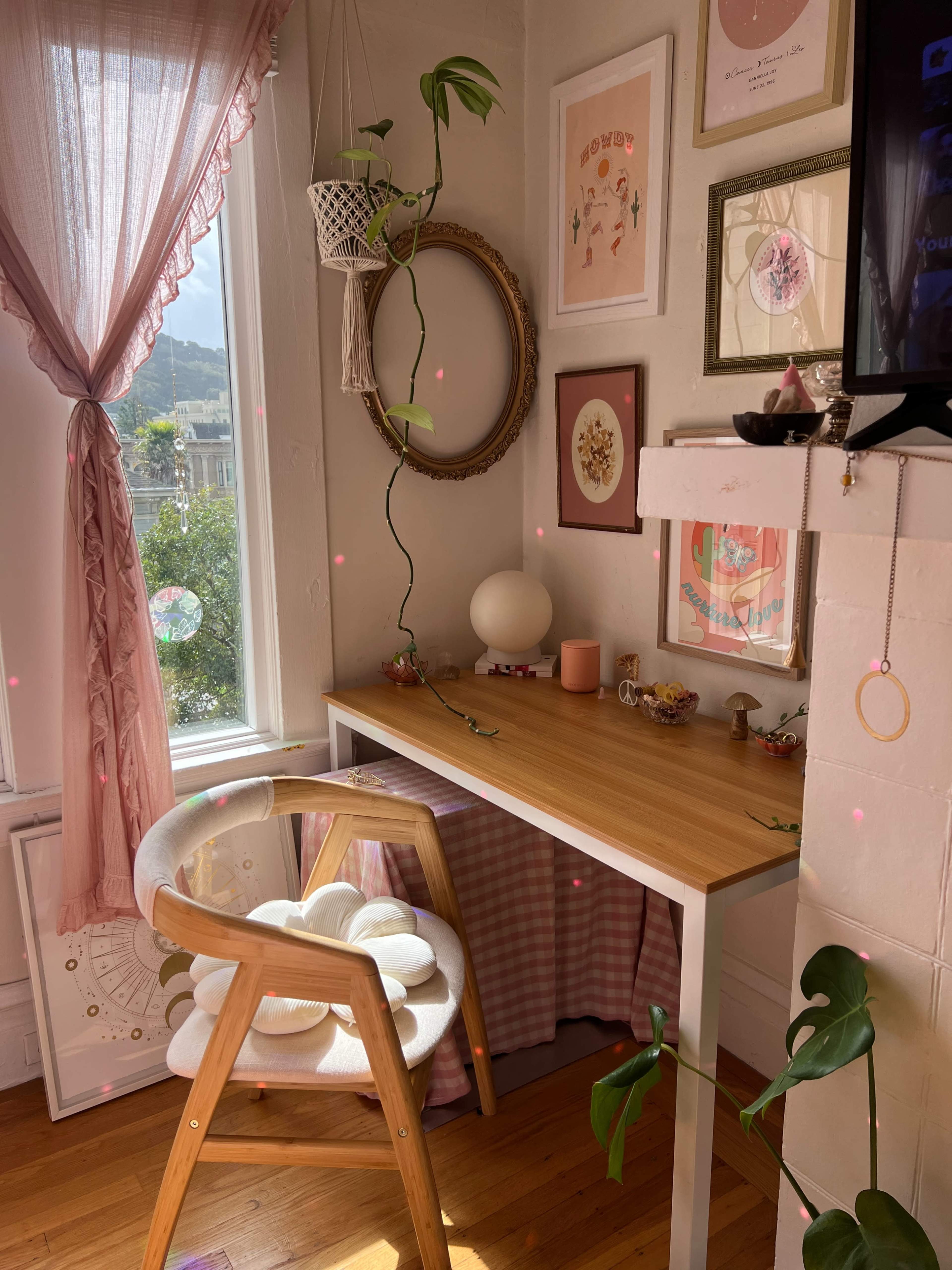 A cozy workspace is set up in a bright corner, featuring a wooden desk, a chair with a floral design, and various decorative art pieces on the walls.