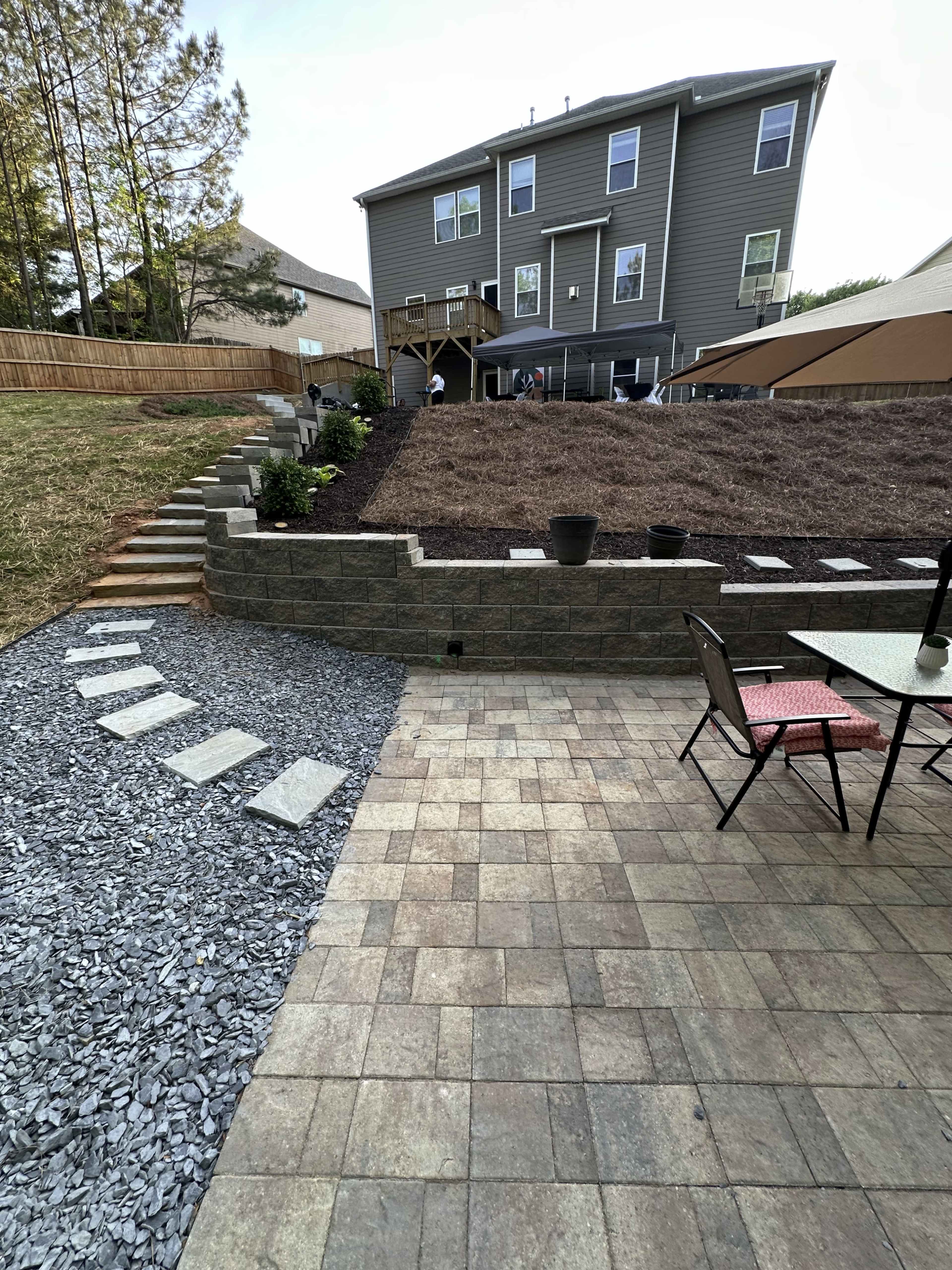 The image shows a backyard with a tiered landscape featuring a stone retaining wall, newly laid mulch, and a patio area with a table and chairs.