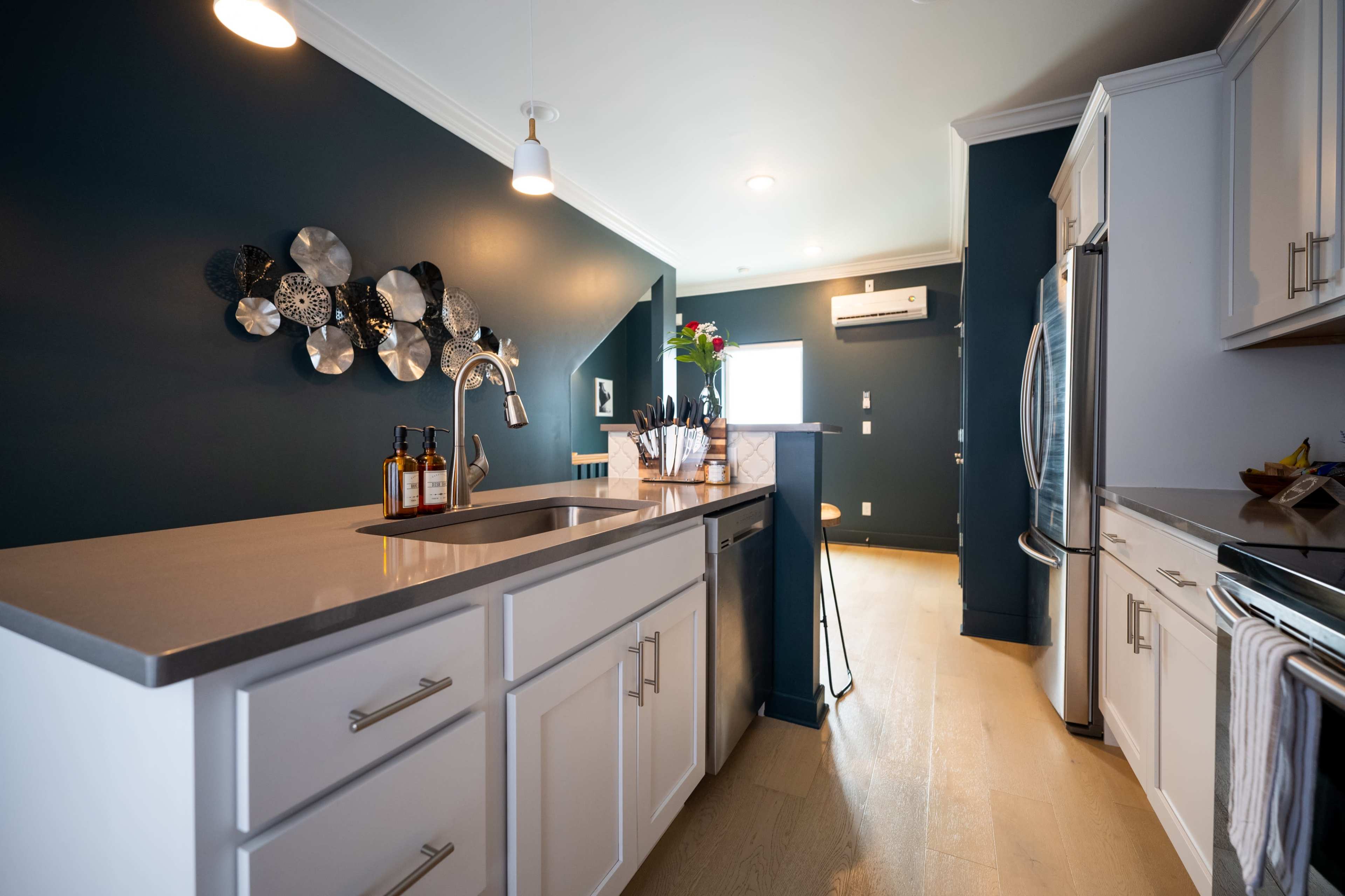 The image shows a modern kitchen with white cabinetry, dark blue walls, and a stainless steel refrigerator, featuring a countertop with kitchen utensils and decorative elements on the wall.