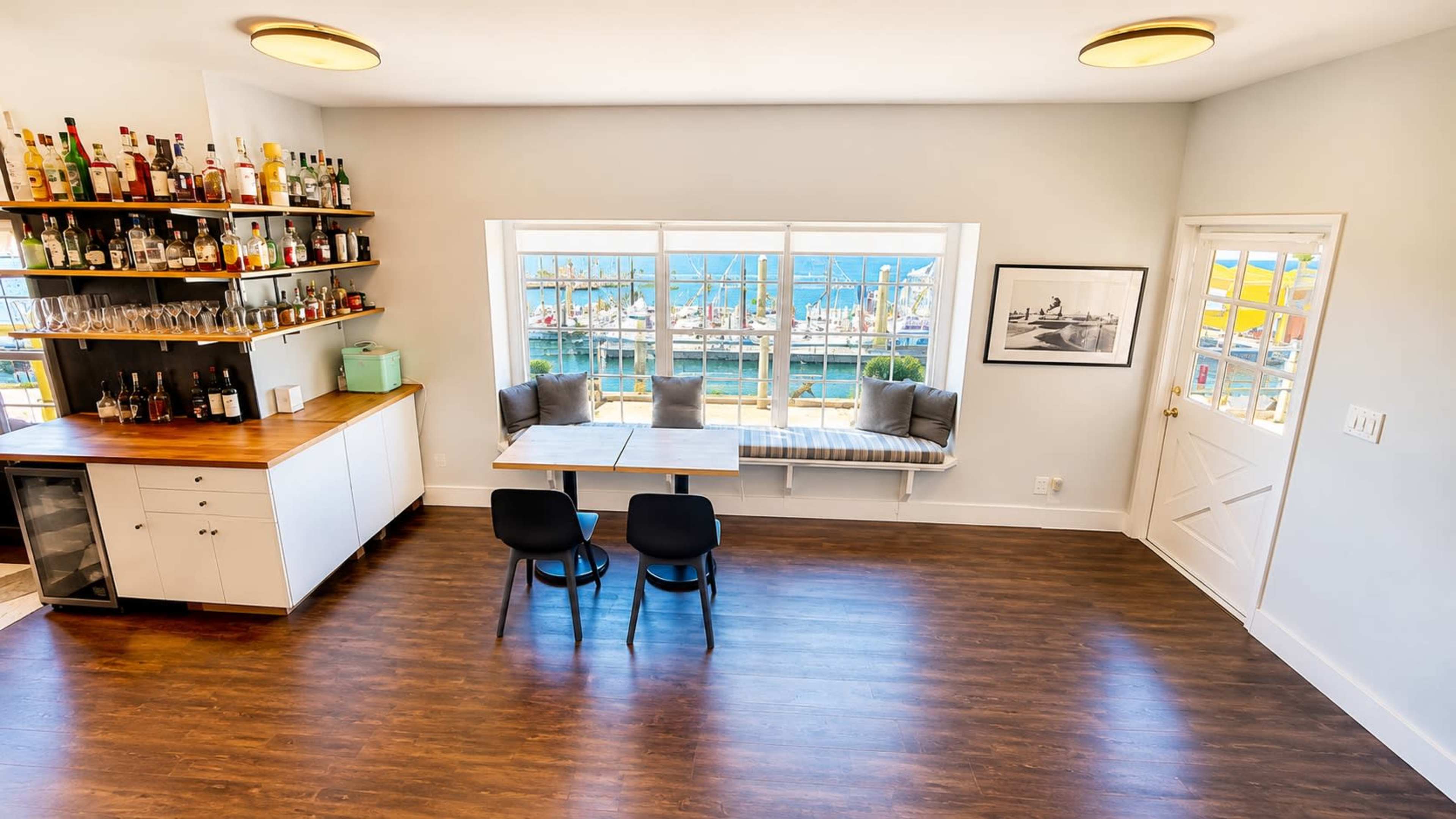 The image shows a modern interior room with a bar area, a dining table with two chairs, and large windows offering a view of a harbor.