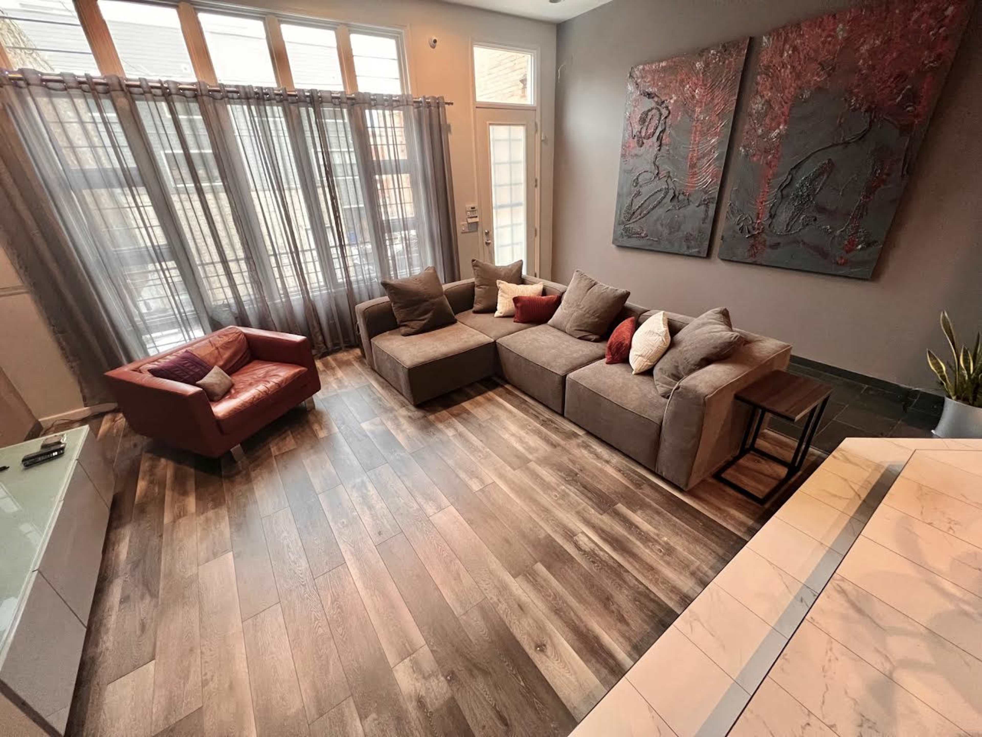 A modern living room features a sectional sofa with various cushions, a small side table, and a single armchair, set against large windows with sheer curtains and two large wall panels.