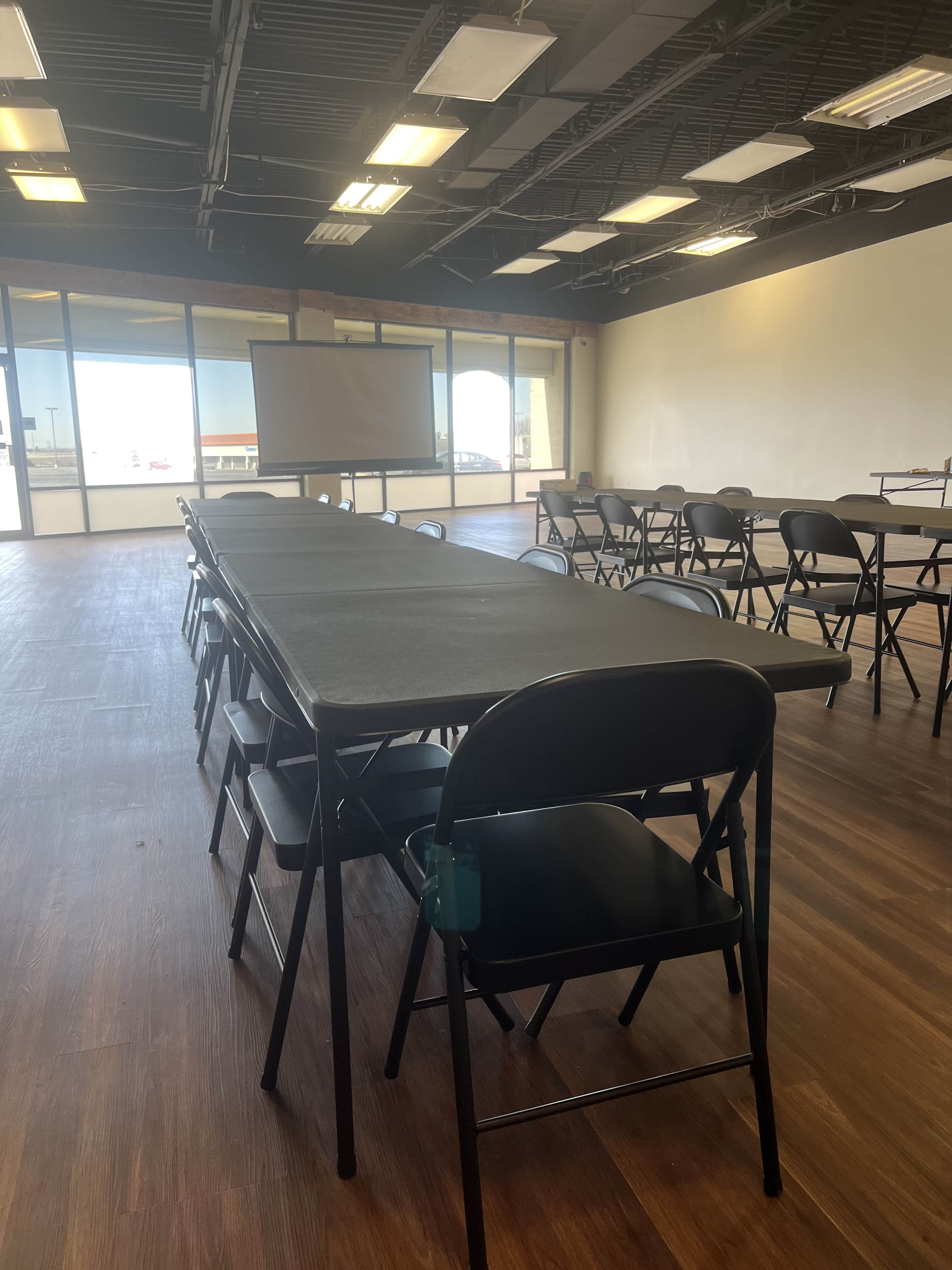 A long table with black folding chairs is set up in a spacious room with large windows and a projector screen at one end.