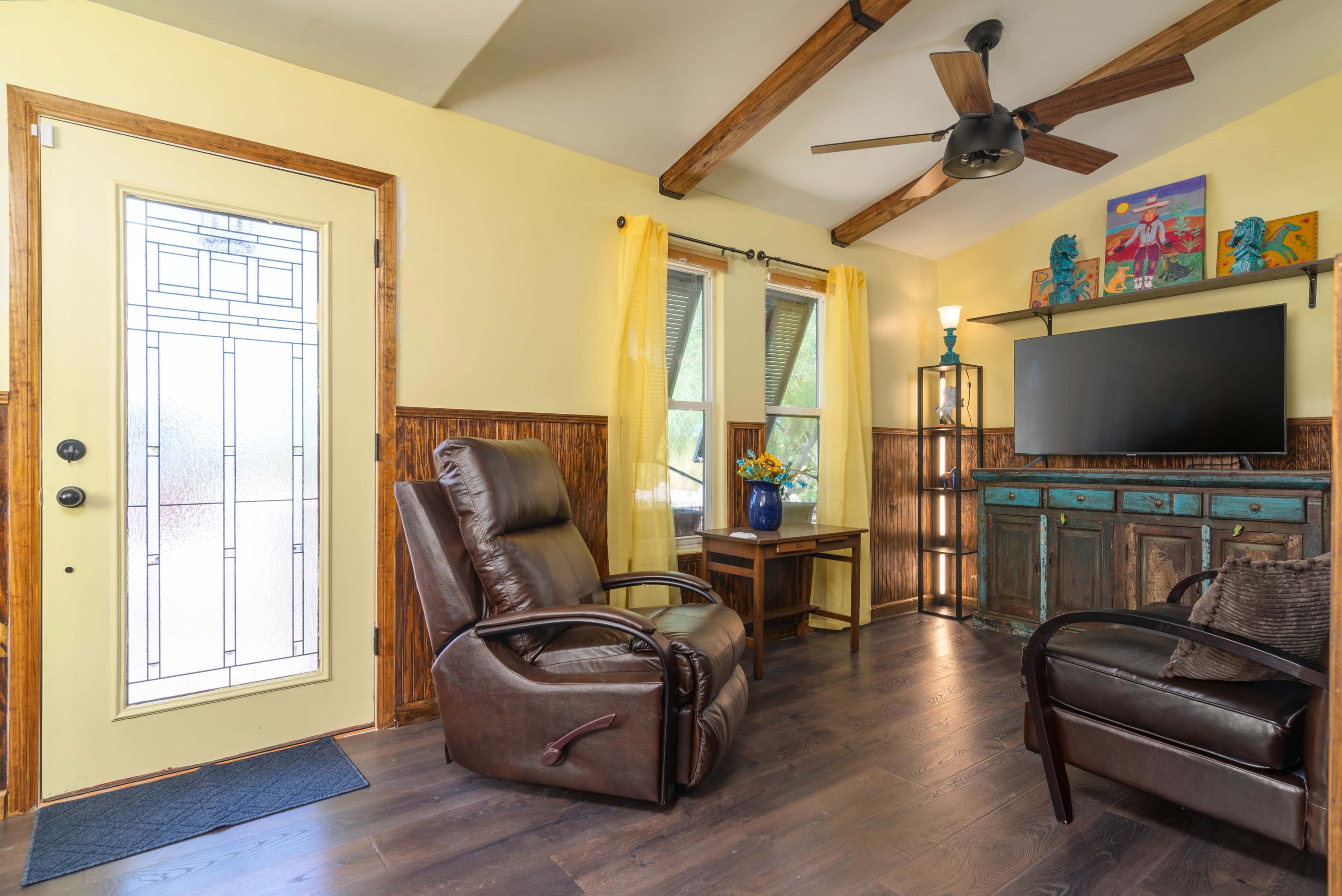The image shows a cozy living room with yellow walls, wooden accents, a door with a decorative glass insert, and a TV mounted on a colorful cabinet.