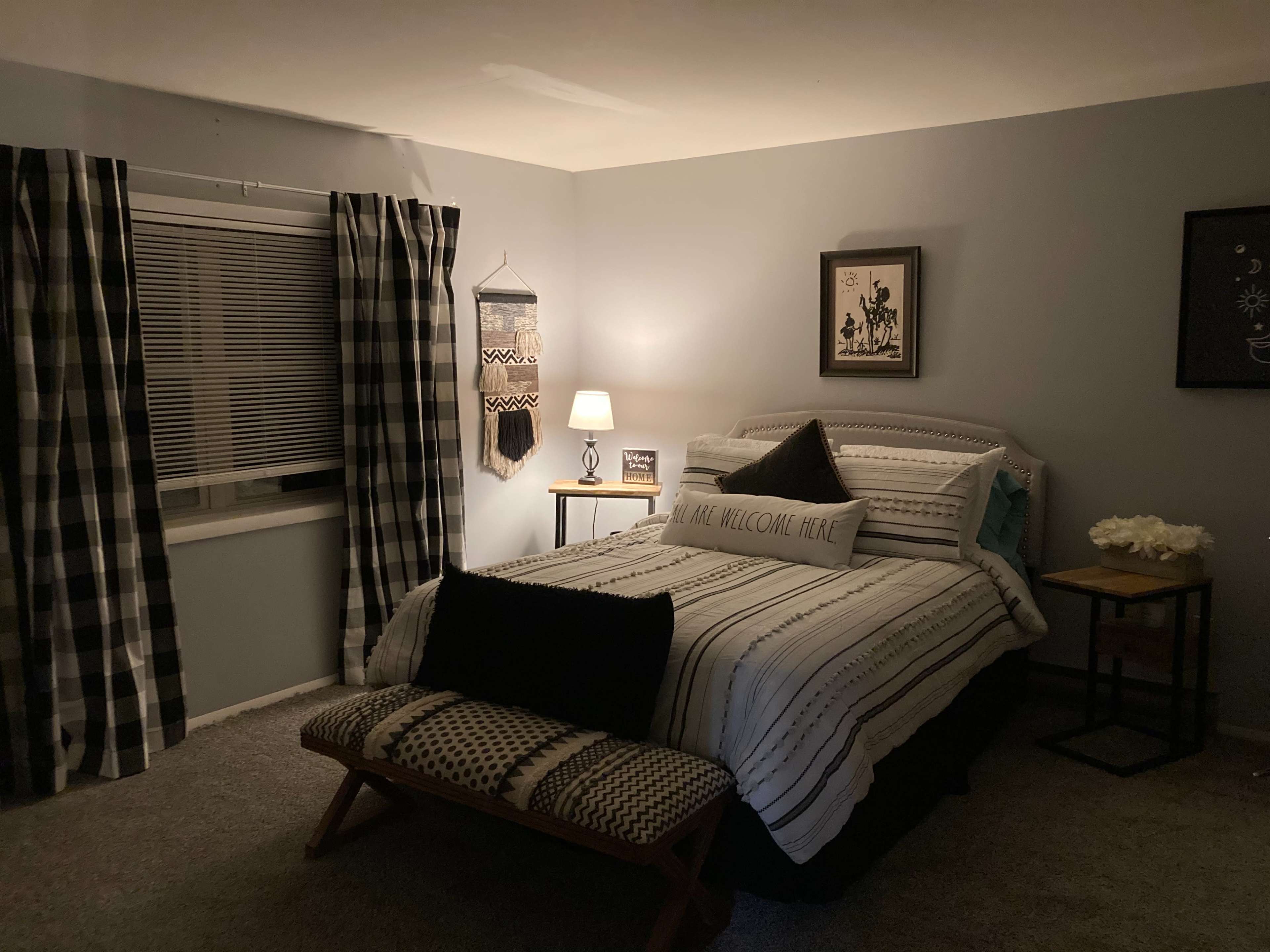 A neatly arranged bedroom features a bed with striped bedding, a bench at the foot, and a lamp on a side table, surrounded by patterned curtains.
