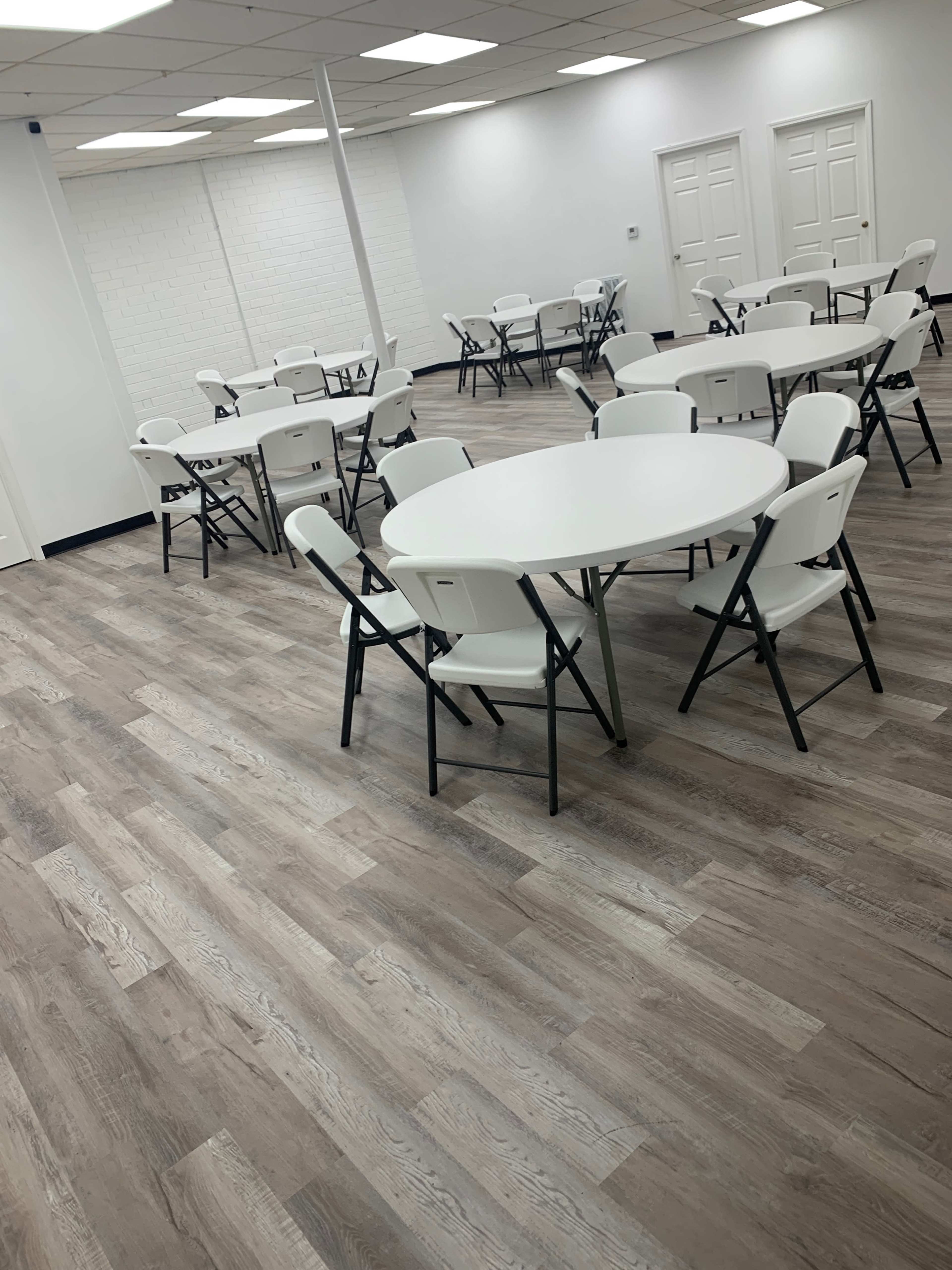 A room with several round tables and white folding chairs arranged neatly on a wooden floor.