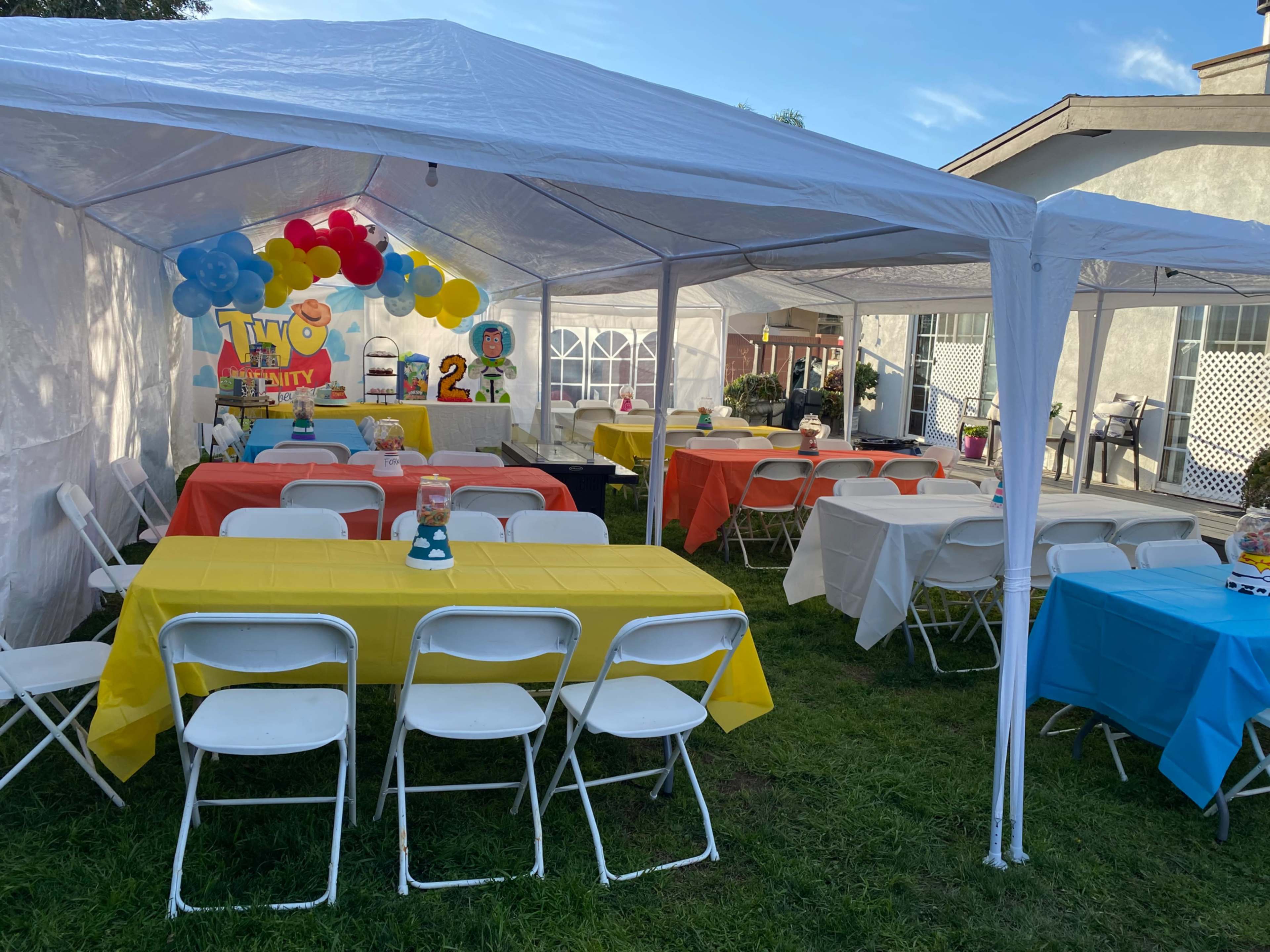 A party tent is set up in a grassy area, featuring colorful tables with decorations, a birthday cake, and balloons.