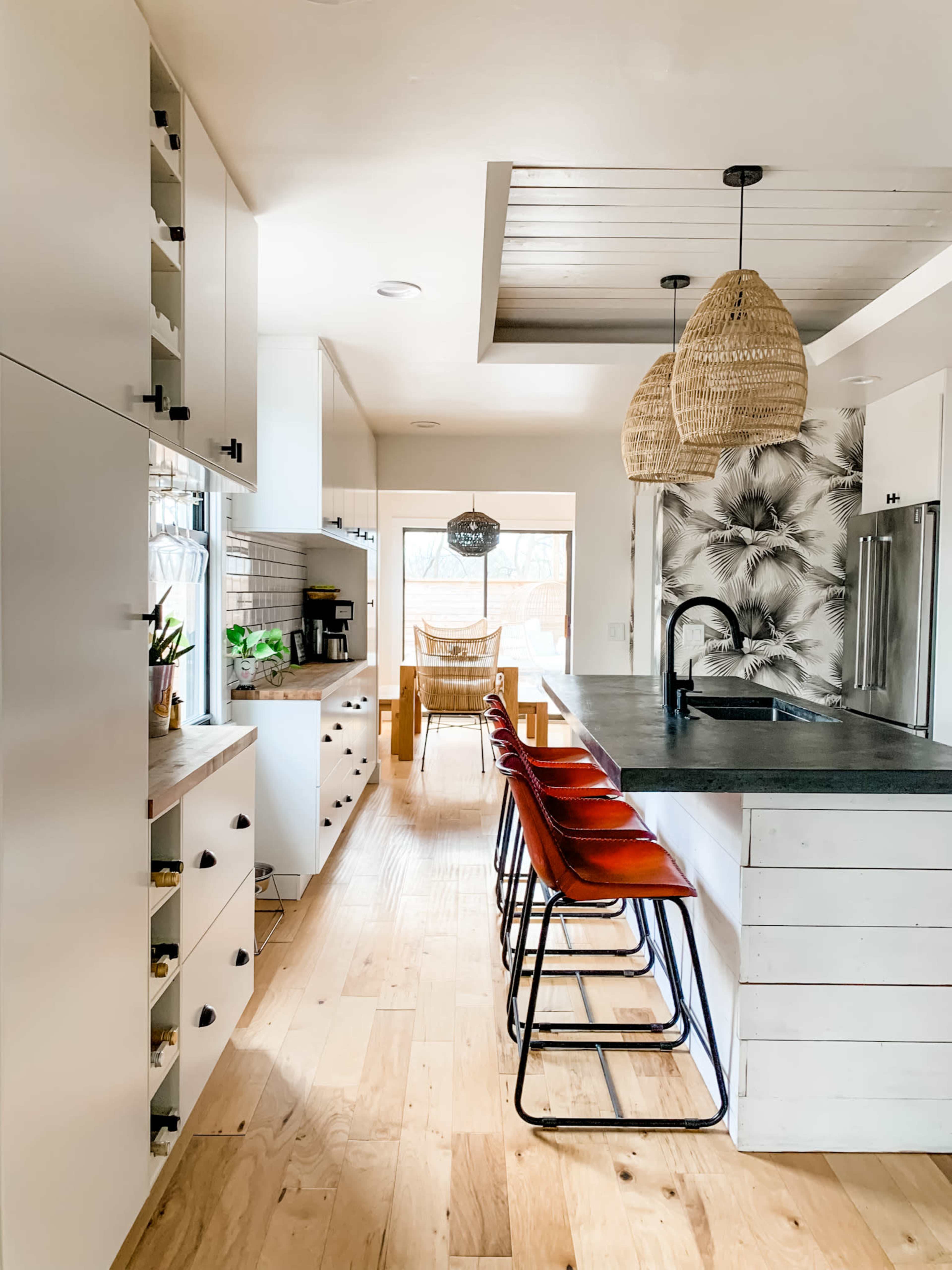 The image shows a modern kitchen with white cabinetry, a concrete countertop, and a dining area featuring a large window and rattan pendant lights.