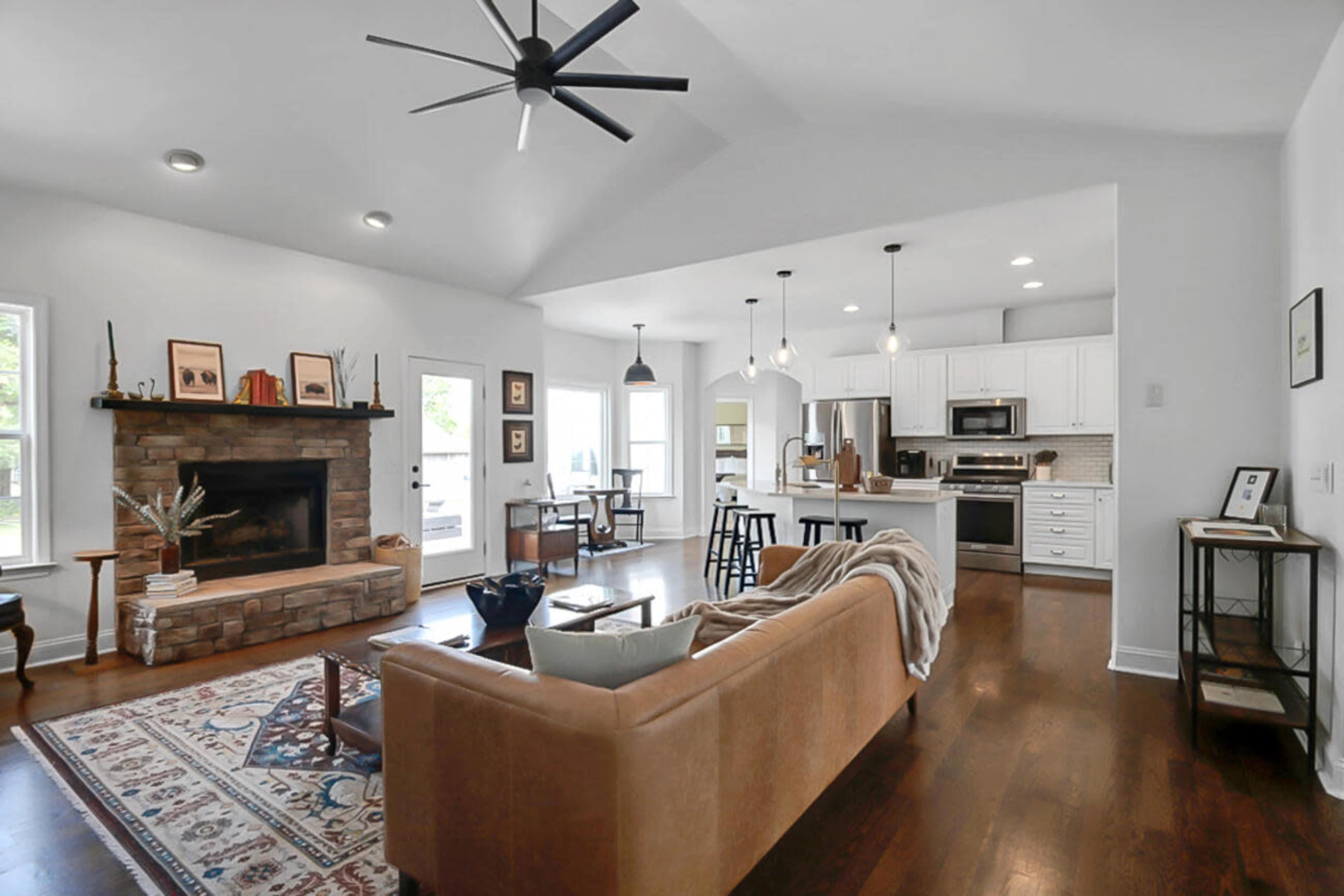 A modern living room with an open layout featuring a stone fireplace, a brown leather sofa, and a view of a kitchen area with bar stools and white cabinets.