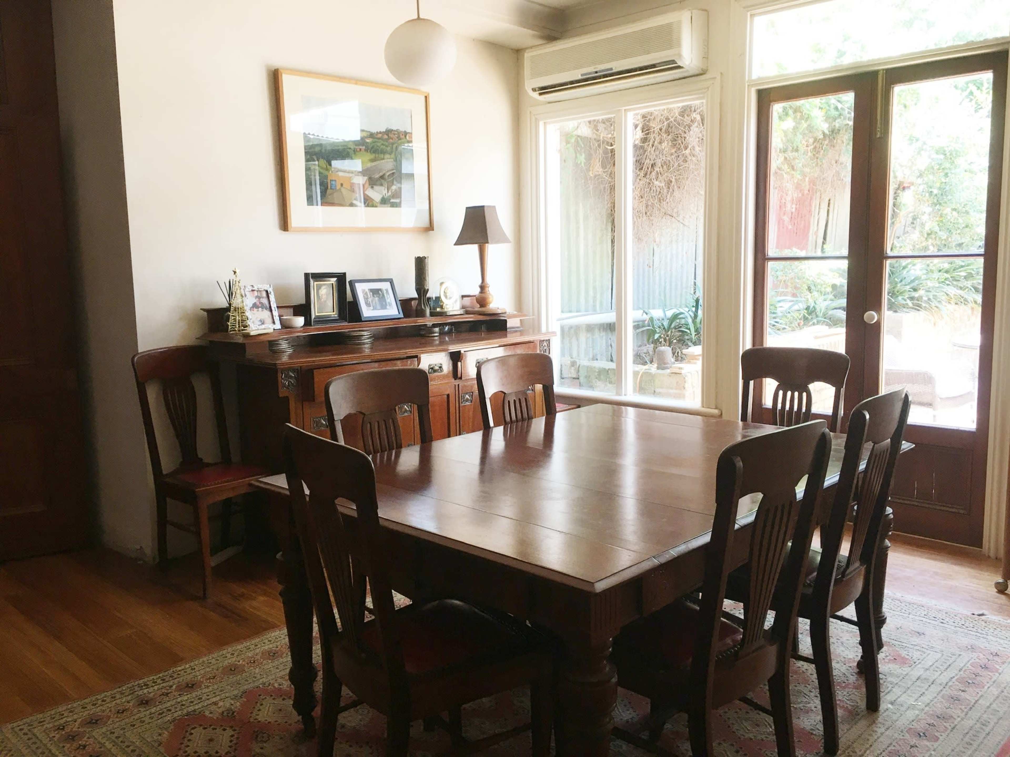 A dining room with a wooden table surrounded by eight chairs, a sideboard with framed photos, and large windows leading to an outdoor area.