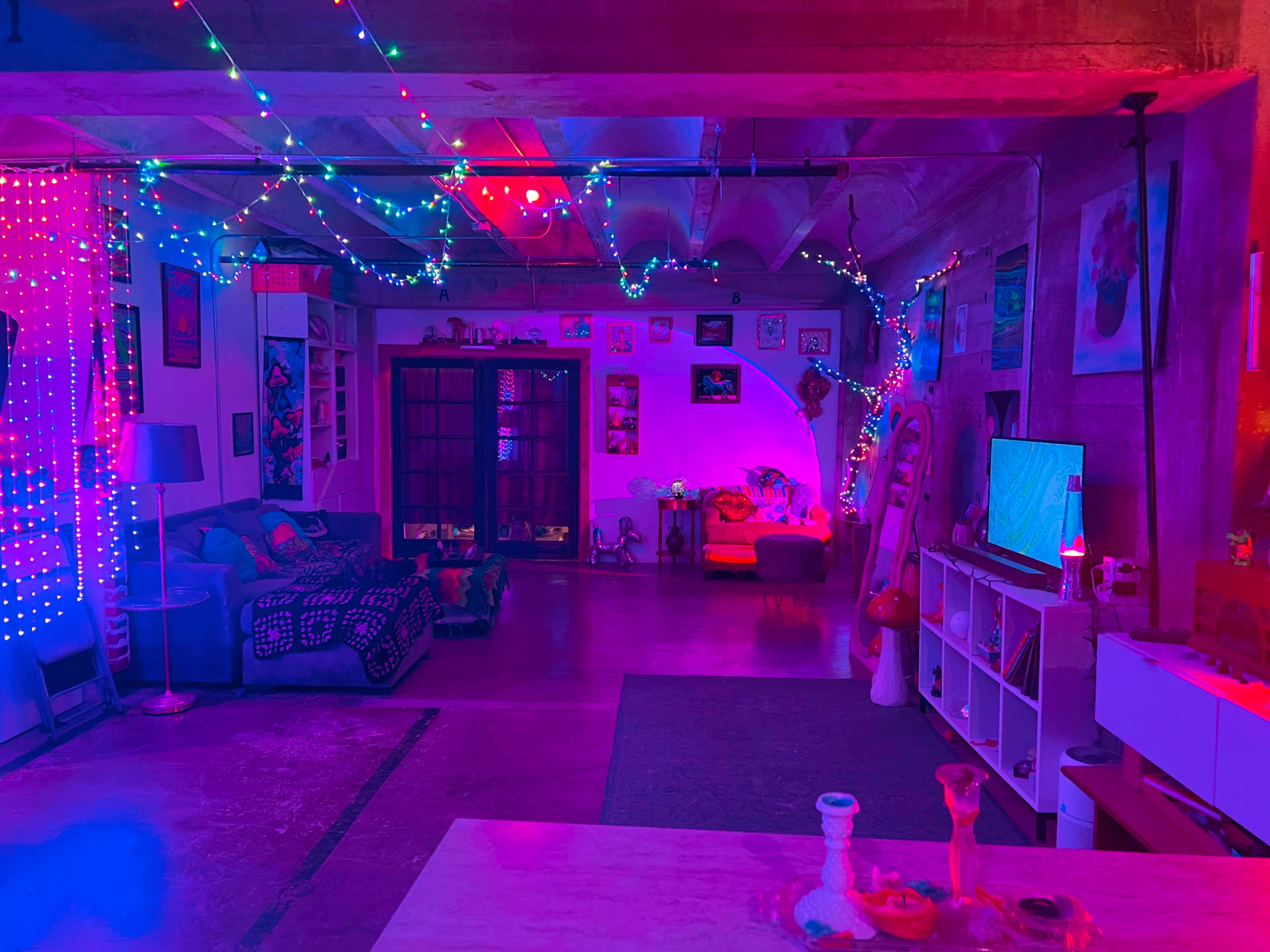 The image shows a vibrant, eclectic living space illuminated with colorful LED lights, featuring a mix of cozy furniture and decorative art on the walls.