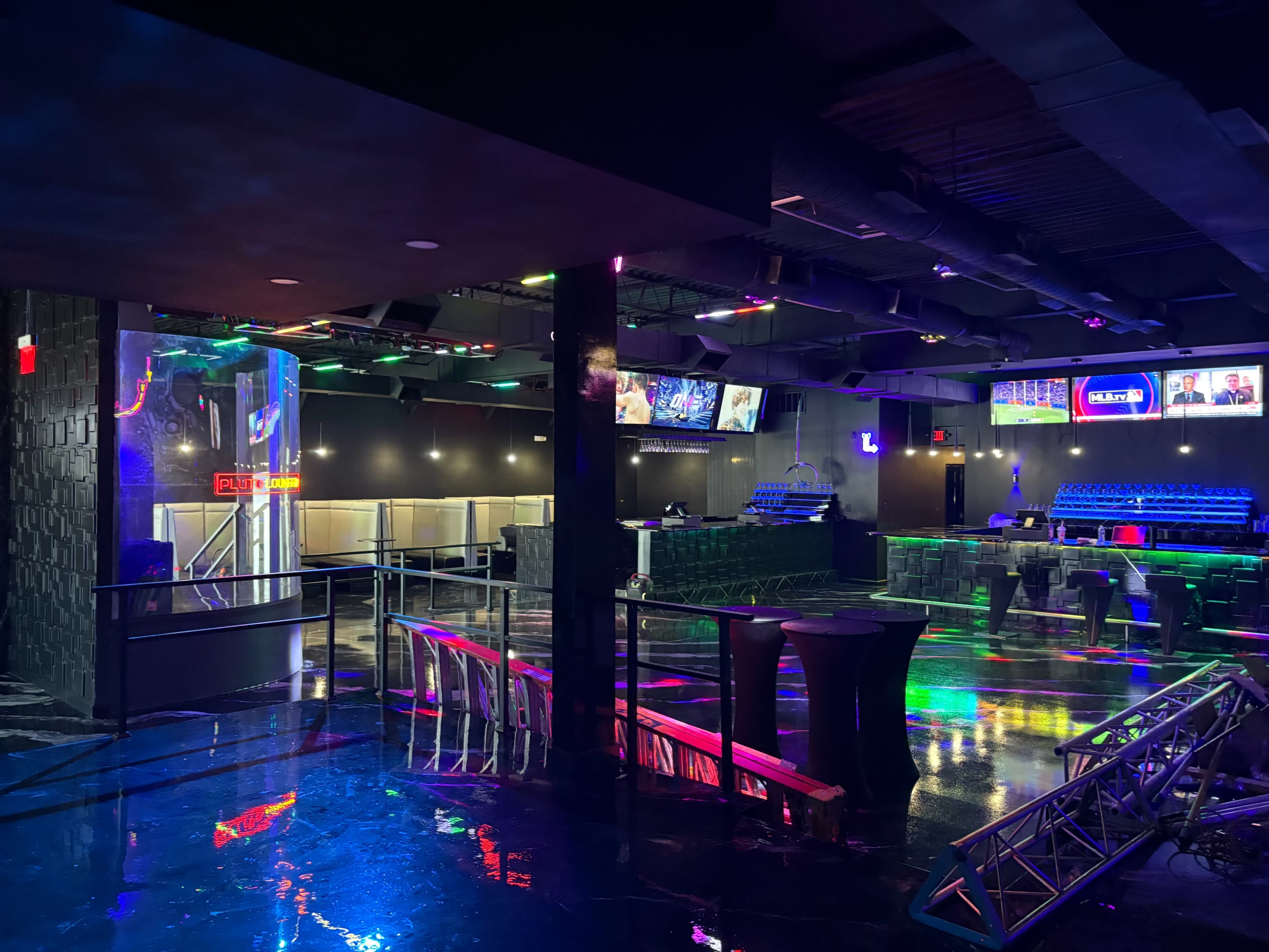 The interior of a dimly lit entertainment venue features colorful lighting, a spacious lounge area with tables, and a bar along one side.