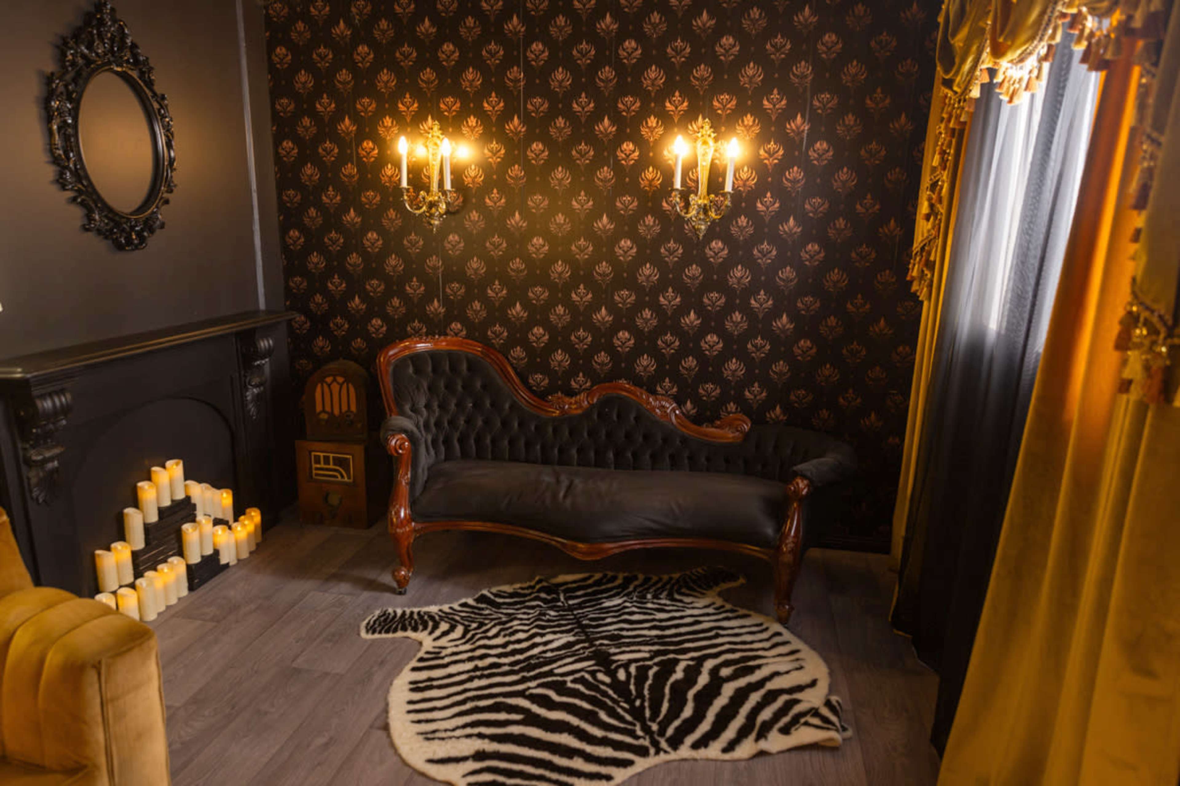 The image shows a vintage-style room with a dark patterned wall, a black leather sofa, a zebra-print rug, wall sconces, and candles arranged by a fireplace.