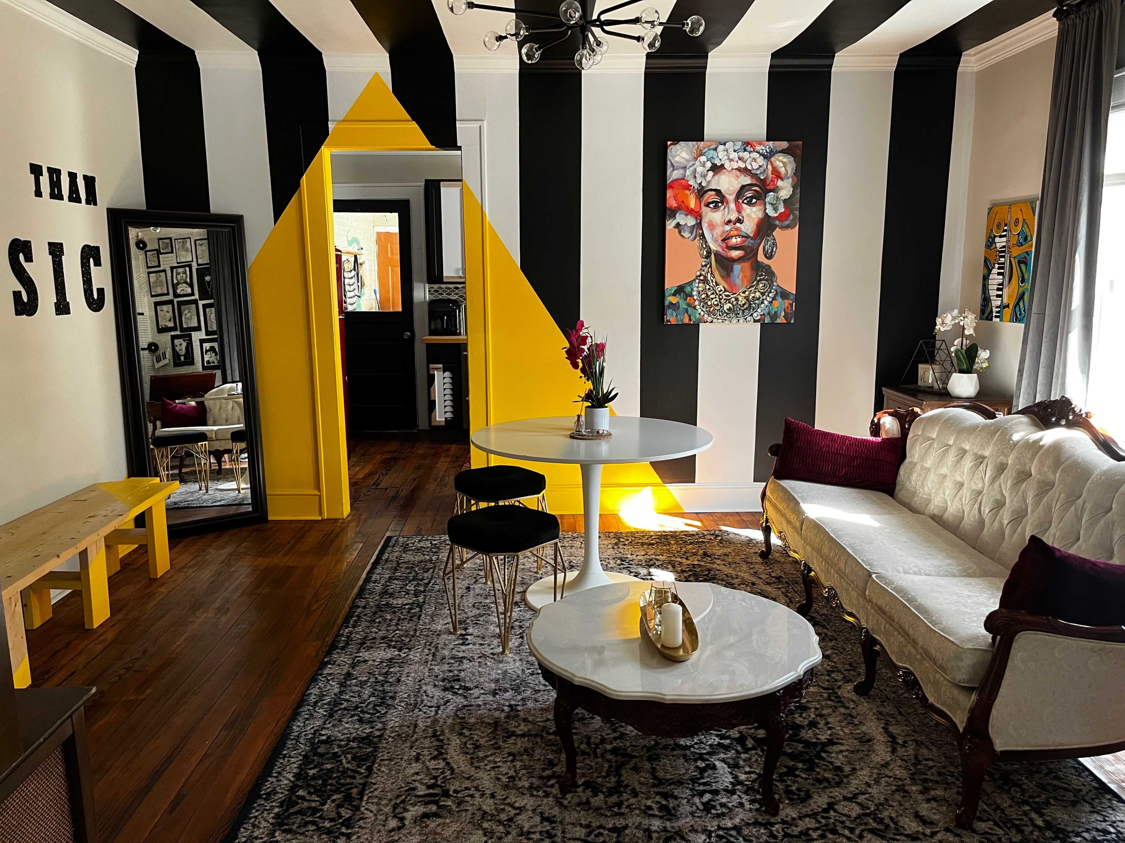 The room features black and white striped walls, a yellow geometric accent wall, a round white dining table, and a vintage sofa, with an artistic portrait displayed above a coffee table.