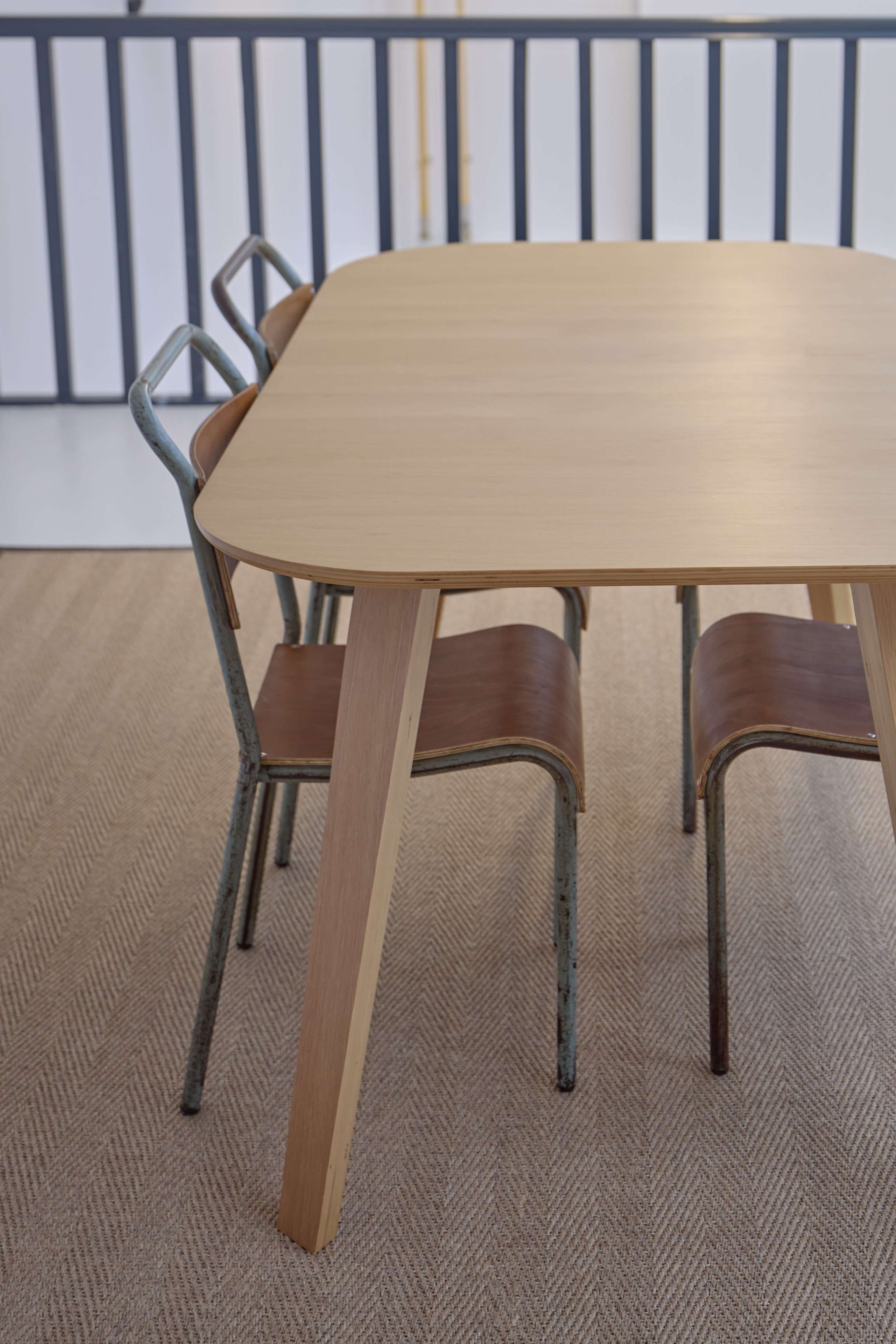 A wooden table with rounded edges is accompanied by two metal chairs with wooden seats, positioned on a textured carpet.