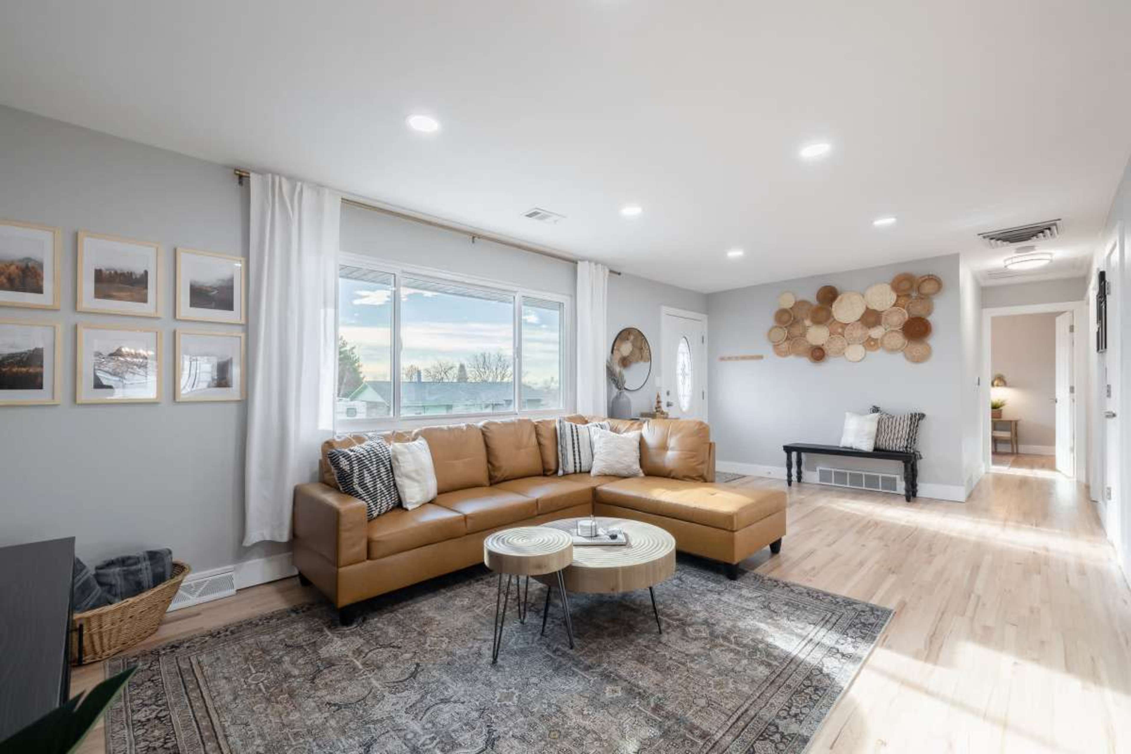 A modern living room features a brown leather sectional sofa, a light-colored coffee table, and wall decorations, with large windows allowing natural light to fill the space.