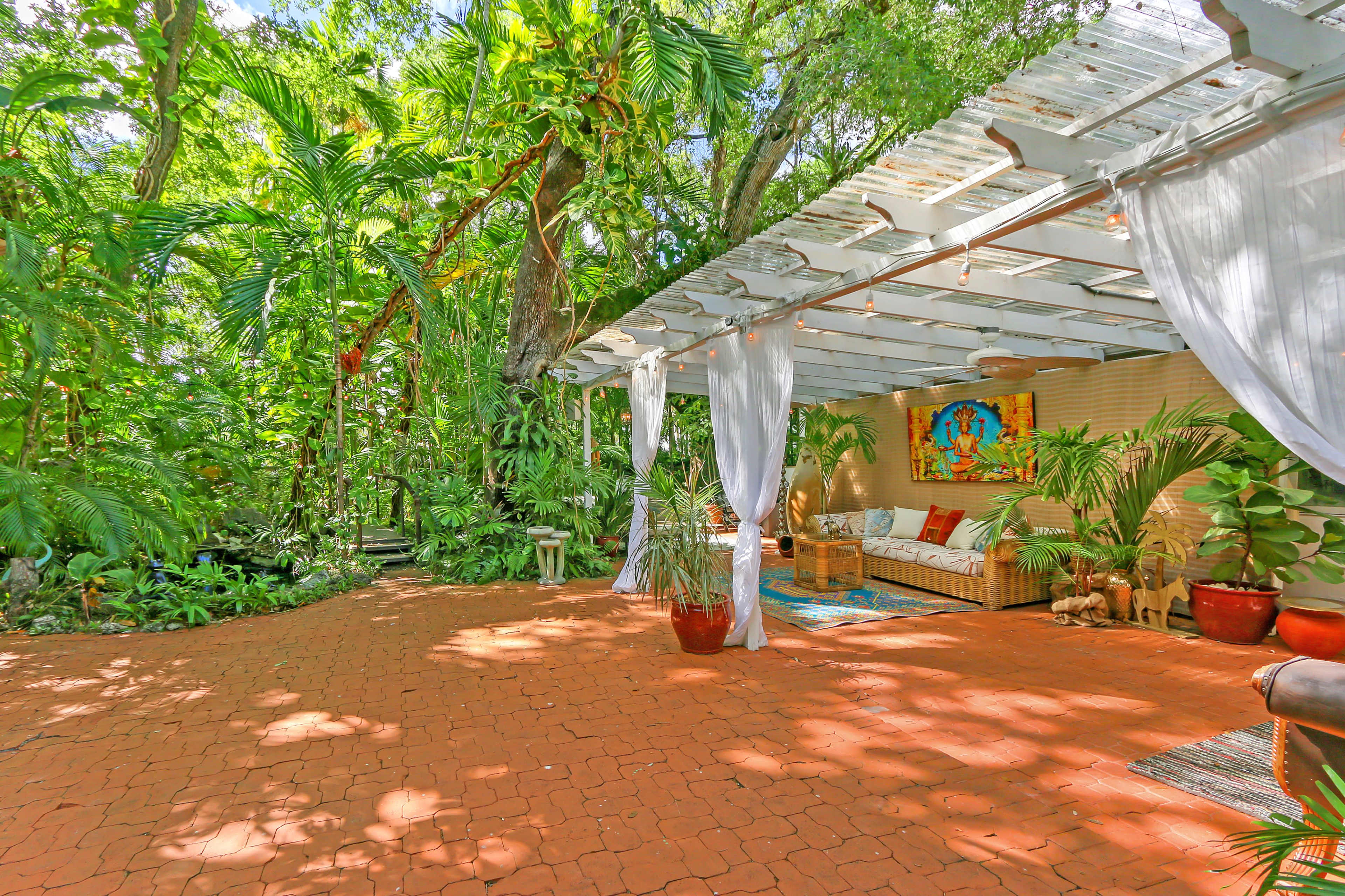 A covered patio with a woven sofa and decorative plants is surrounded by lush greenery.