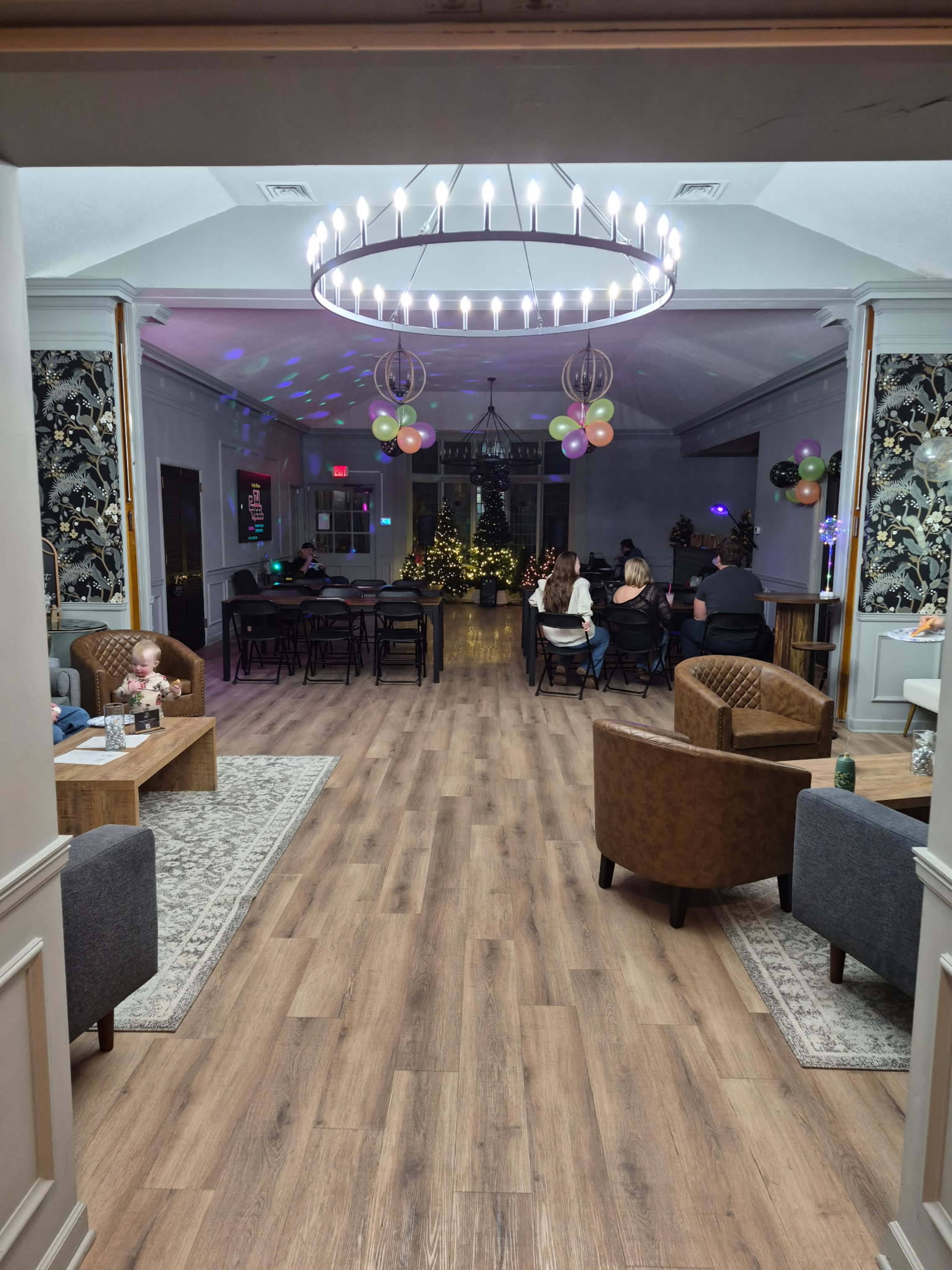The image shows a spacious event room with a seating area and decorative lighting, featuring chairs arranged toward the back and balloons in various colors adorned around the space.