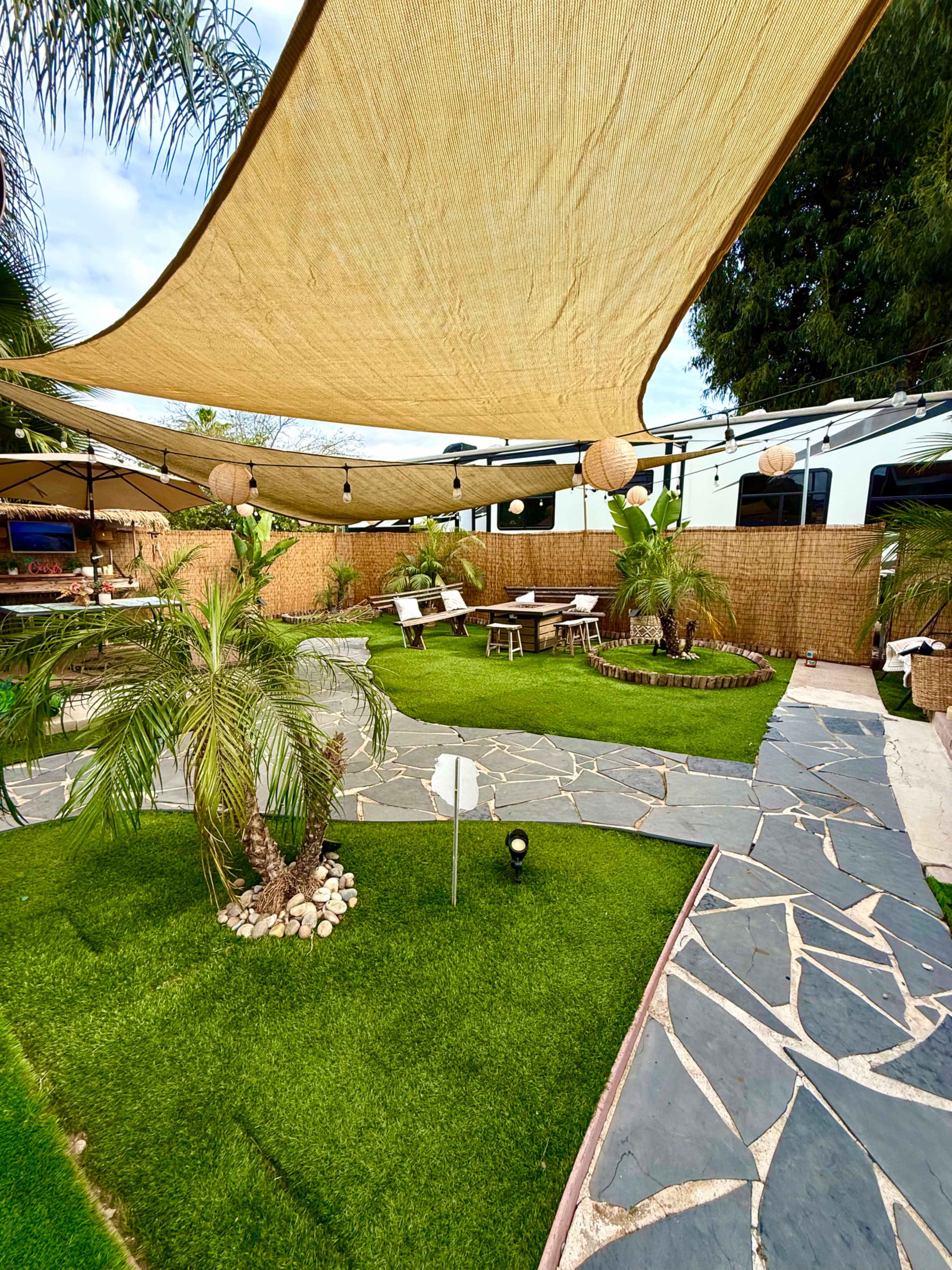 The image shows a landscaped outdoor area with a stone pathway, grass, palm trees, and shaded seating arrangements under a large canopy.