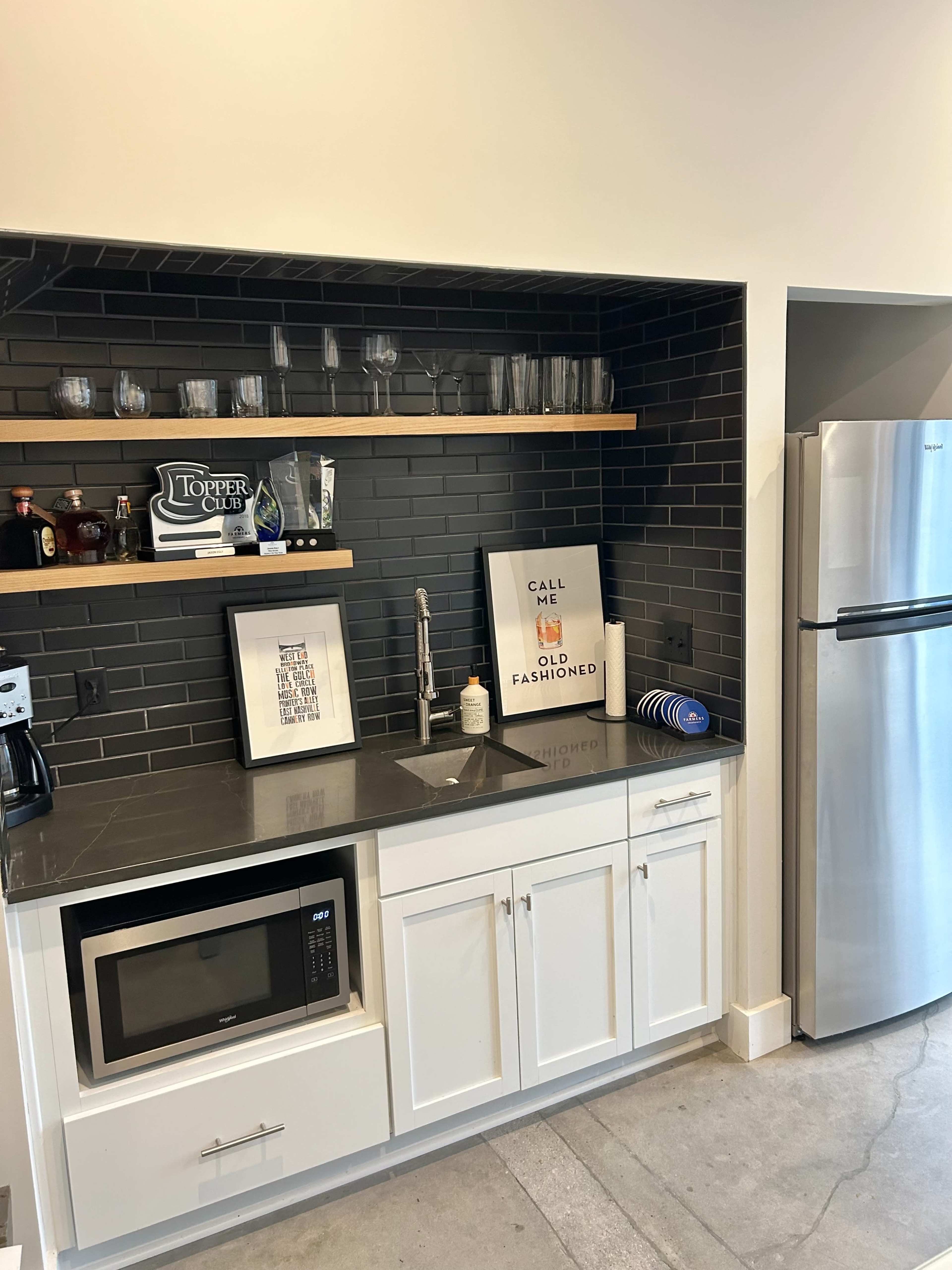The image shows a modern kitchenette featuring a dark tiled backsplash, a stainless steel refrigerator, a microwave, a sink with a faucet, and shelves displaying glassware and decor.