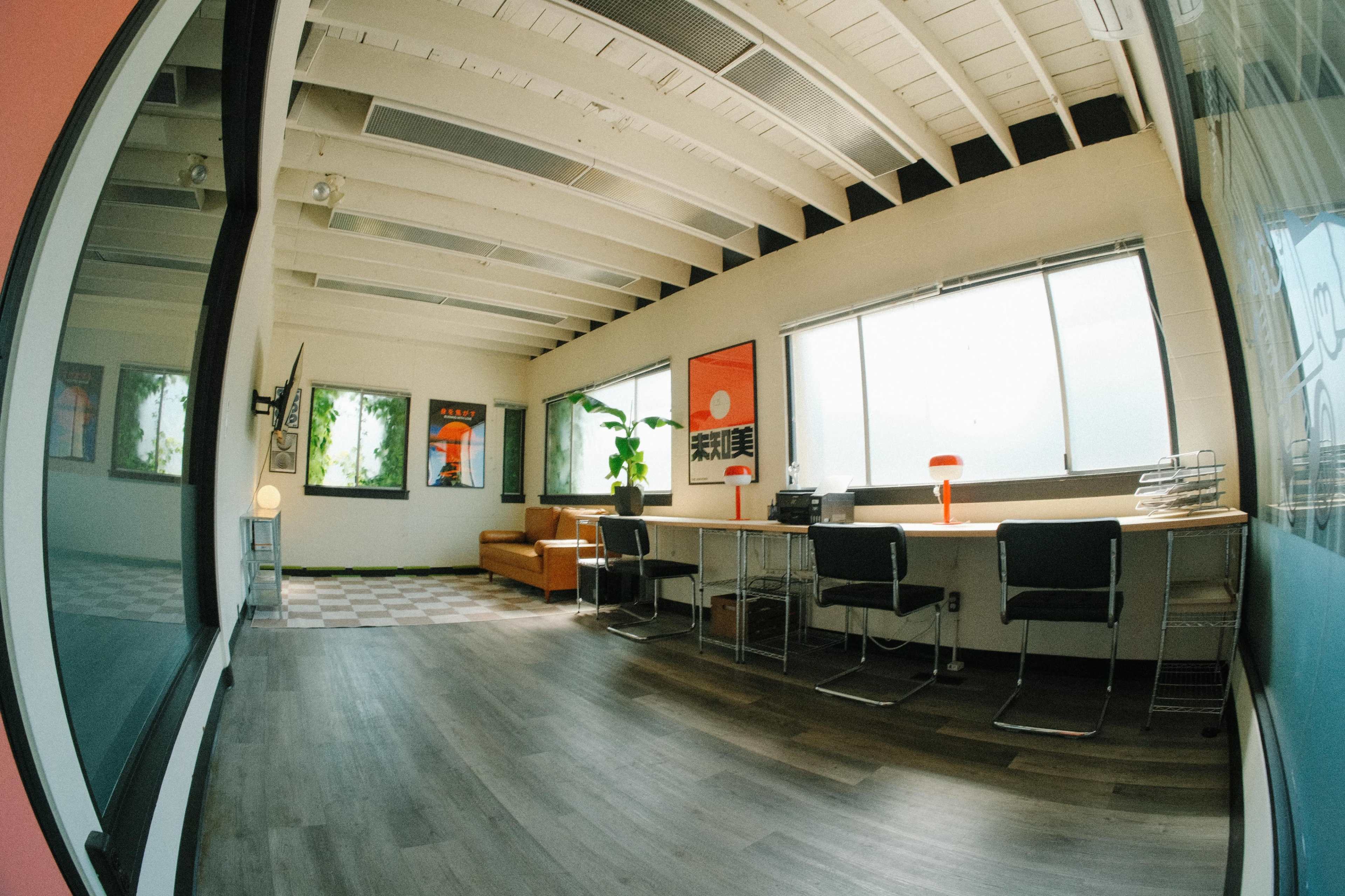 The image shows a spacious office with wooden beams, large windows, and a seating area, featuring minimalist furniture and artwork on the walls.