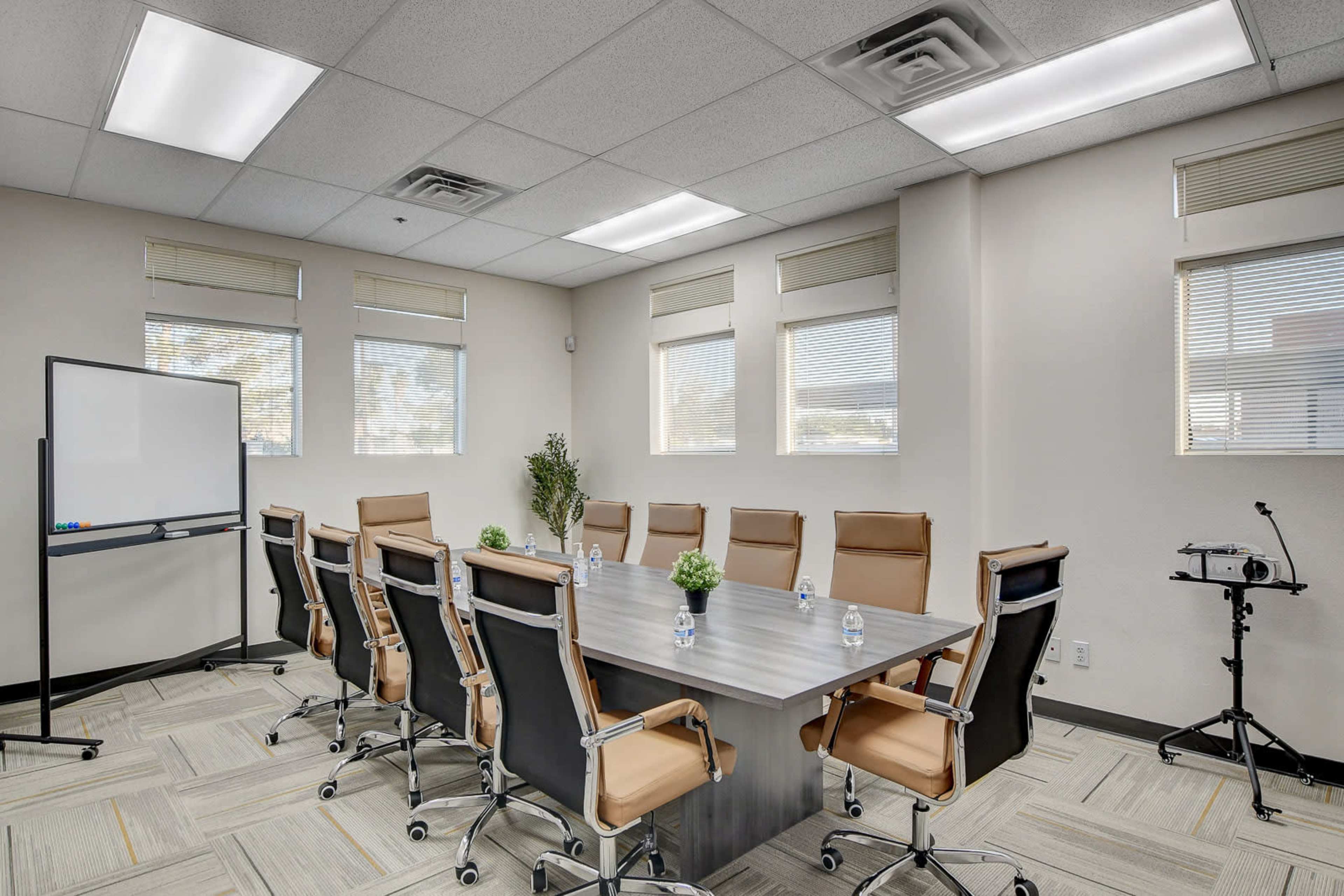 The image shows a conference room with a large table surrounded by several ergonomic chairs, a whiteboard, and a small plant near the window.