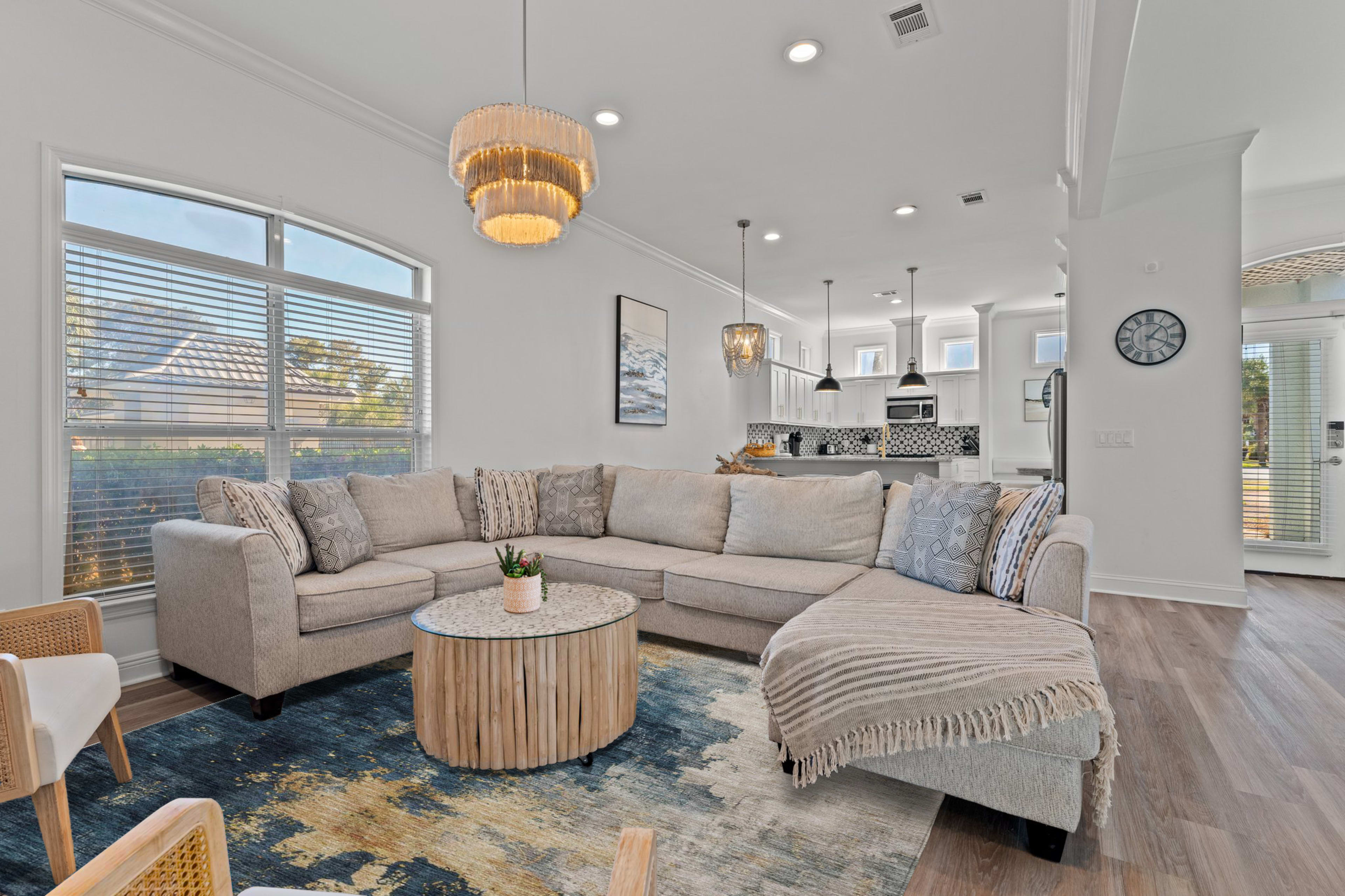 The living room features a large sectional sofa, a round coffee table made of wood, and large windows that allow natural light to illuminate the space.