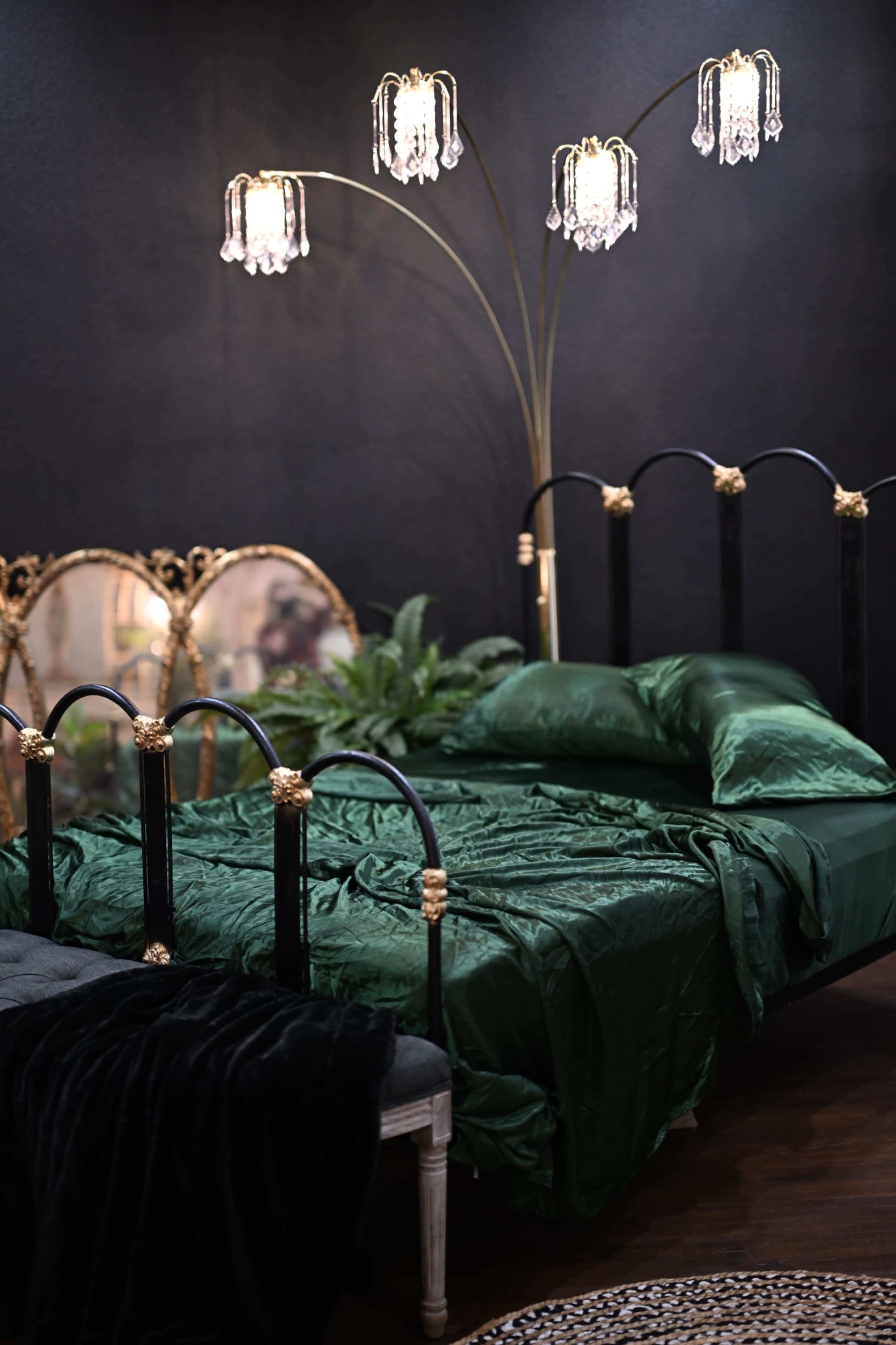 A dark room with a black metal bed covered in a green satin duvet, illuminated by an ornate floor lamp with crystal accents and accompanied by vintage mirrors and decorative plants.