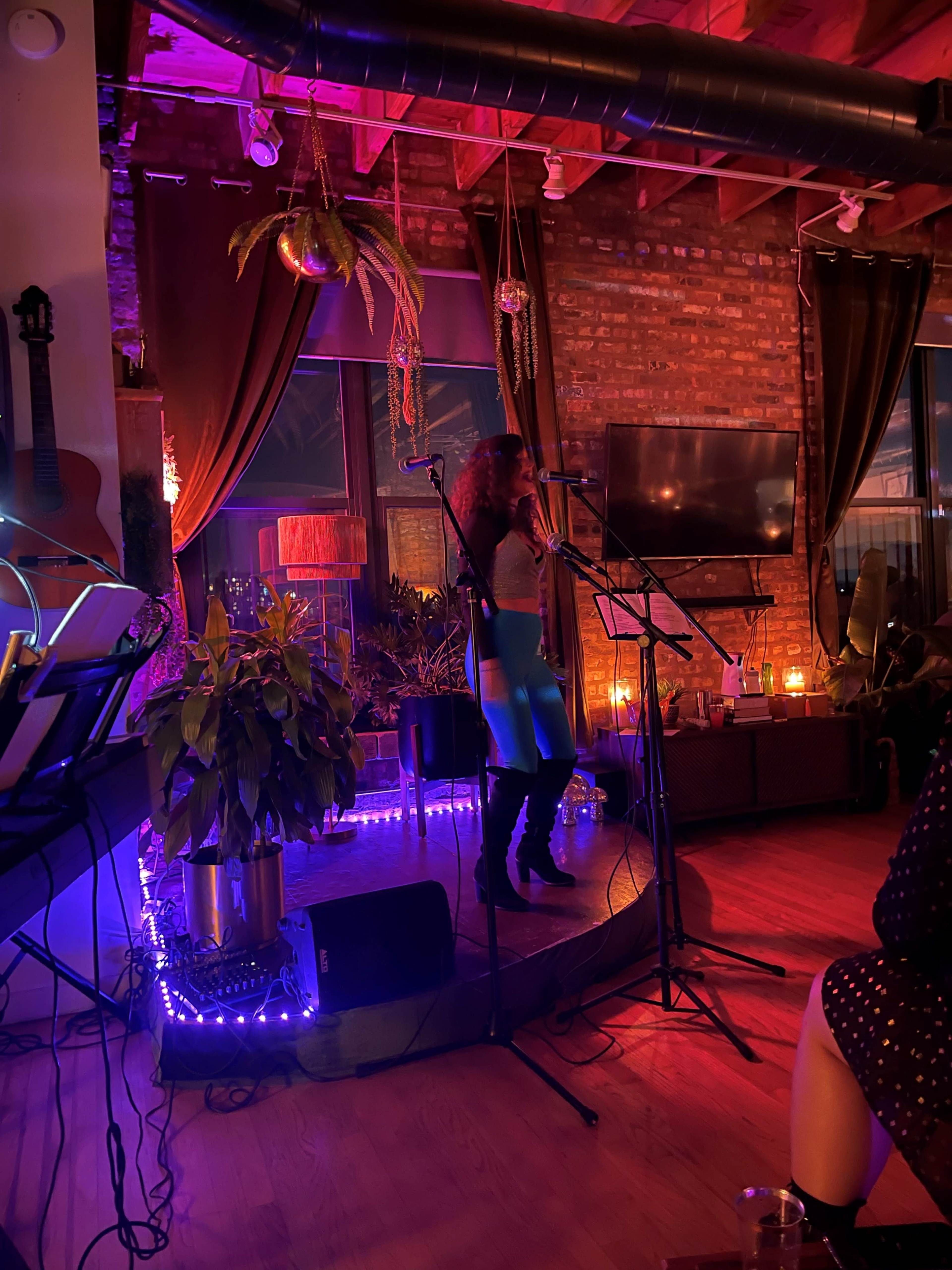 A performer stands on a stage in a dimly lit venue, surrounded by plants and decorative lights, while microphones and musical instruments are set up nearby.