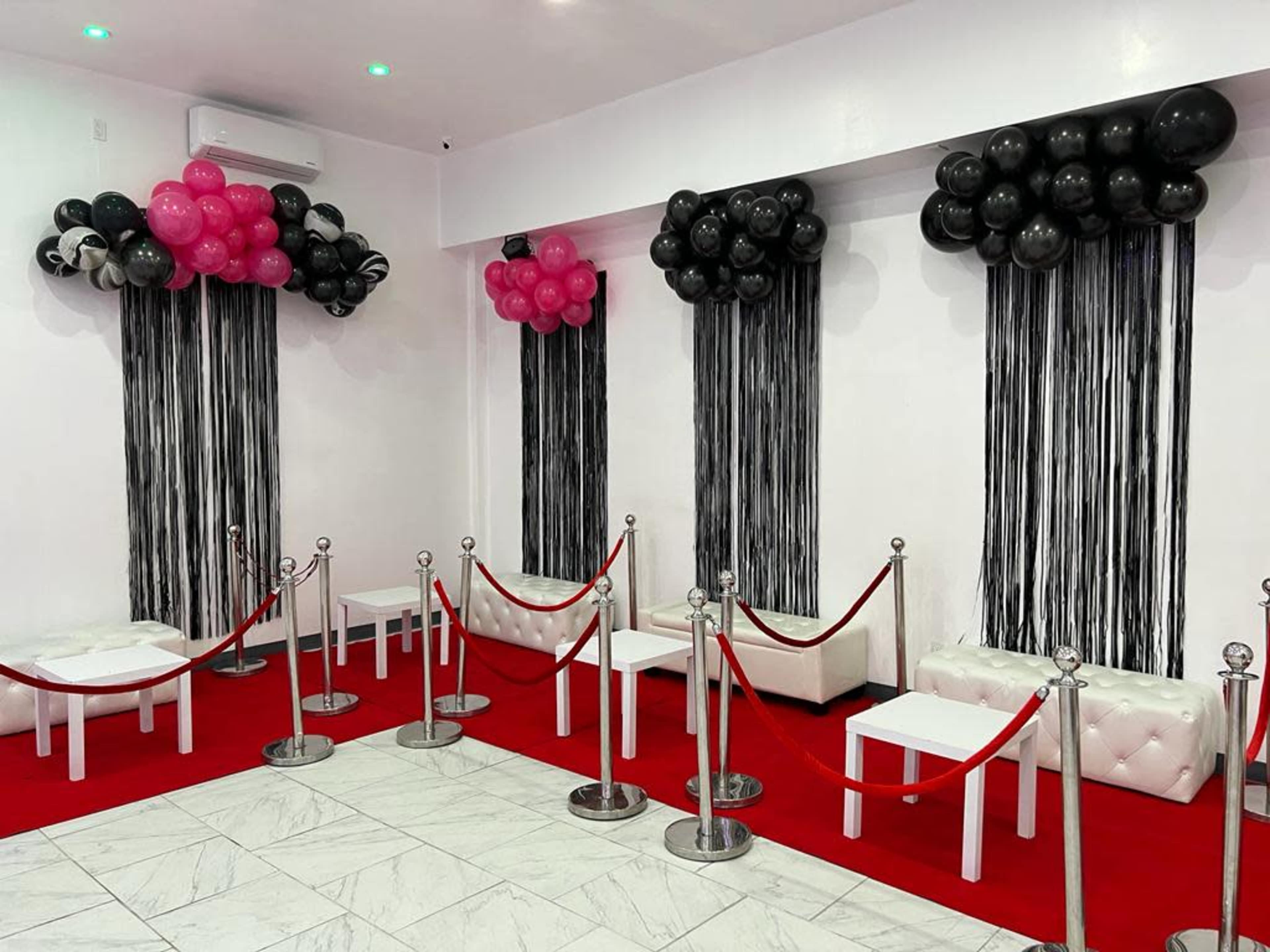 A decorated event space featuring white leather benches, red carpet, and balloon arrangements in black and pink above vertical black streamers.